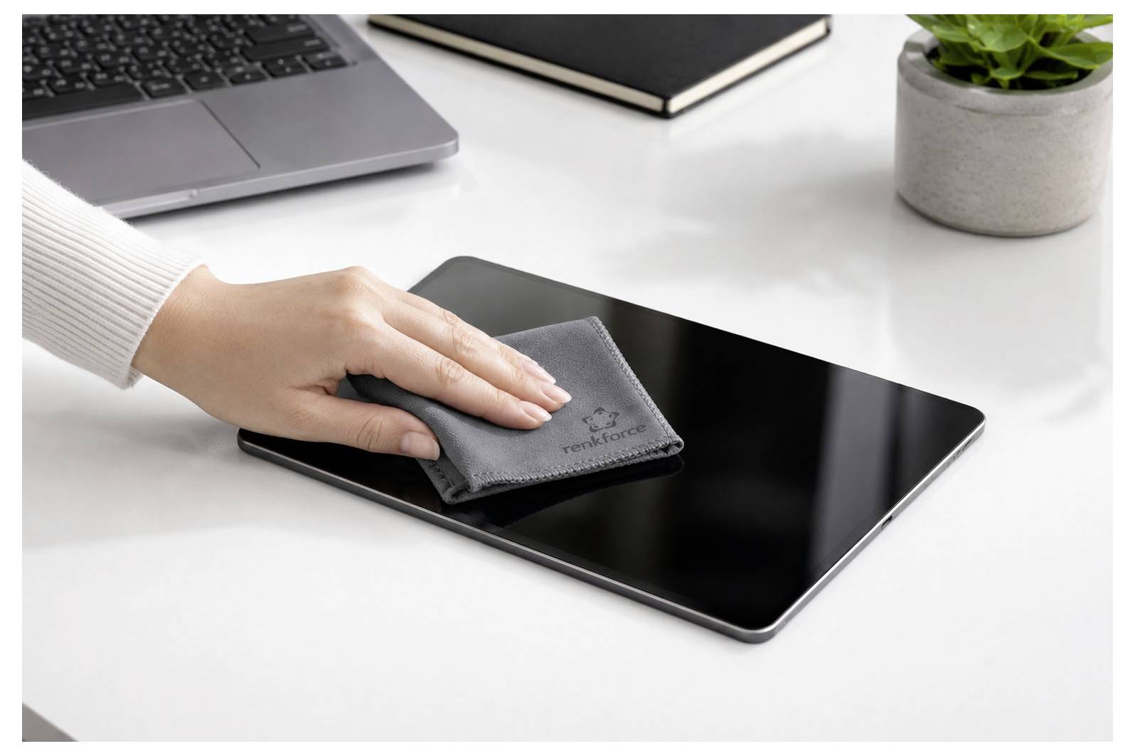 Essuyage à la main d'un écran de tablette noire avec un chiffon gris sur un bureau à côté d'un ordinateur portable et d'une plante.