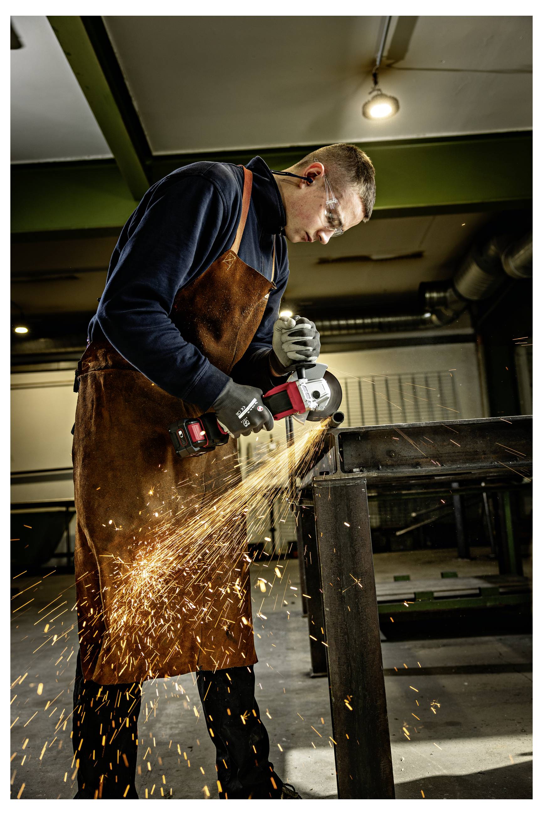 Homme en tablier et gants utilisant une meuleuse d'angle sur une table métallique, envoyant des étincelles brillantes dans un atelier.