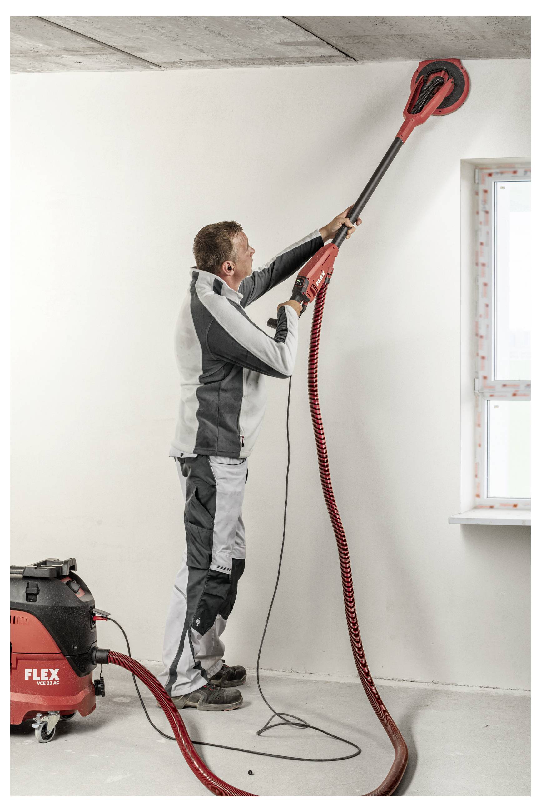 Ouvrier utilisant une ponceuse électrique à long manche pour poncer un plafond dans une pièce en rénovation, avec un extracteur de poussière à côté de lui.