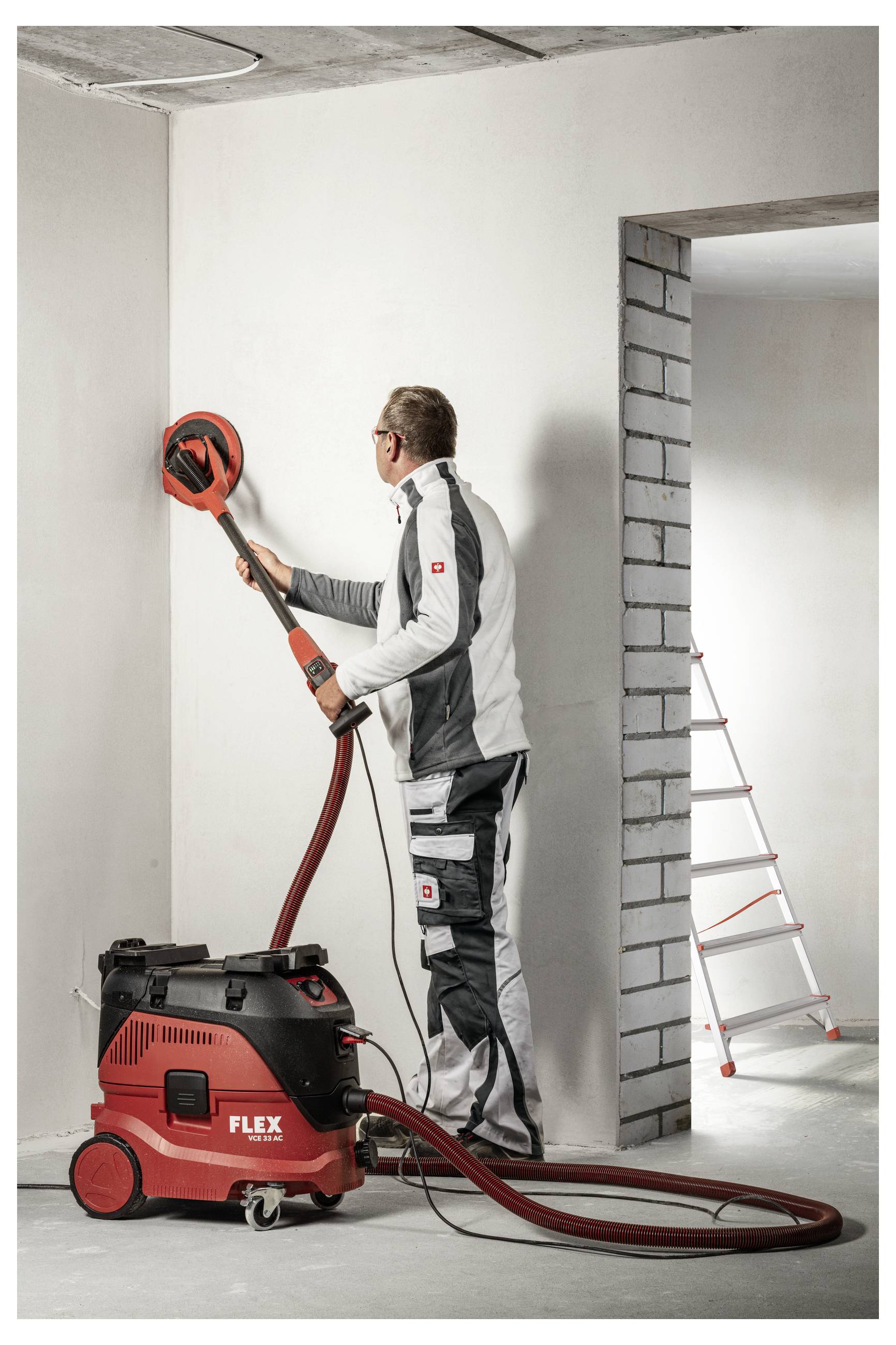 Travailleur utilisant un ponceuse murale rouge attachée à un aspirateur dans une pièce inachevée, avec une échelle visible par la porte.