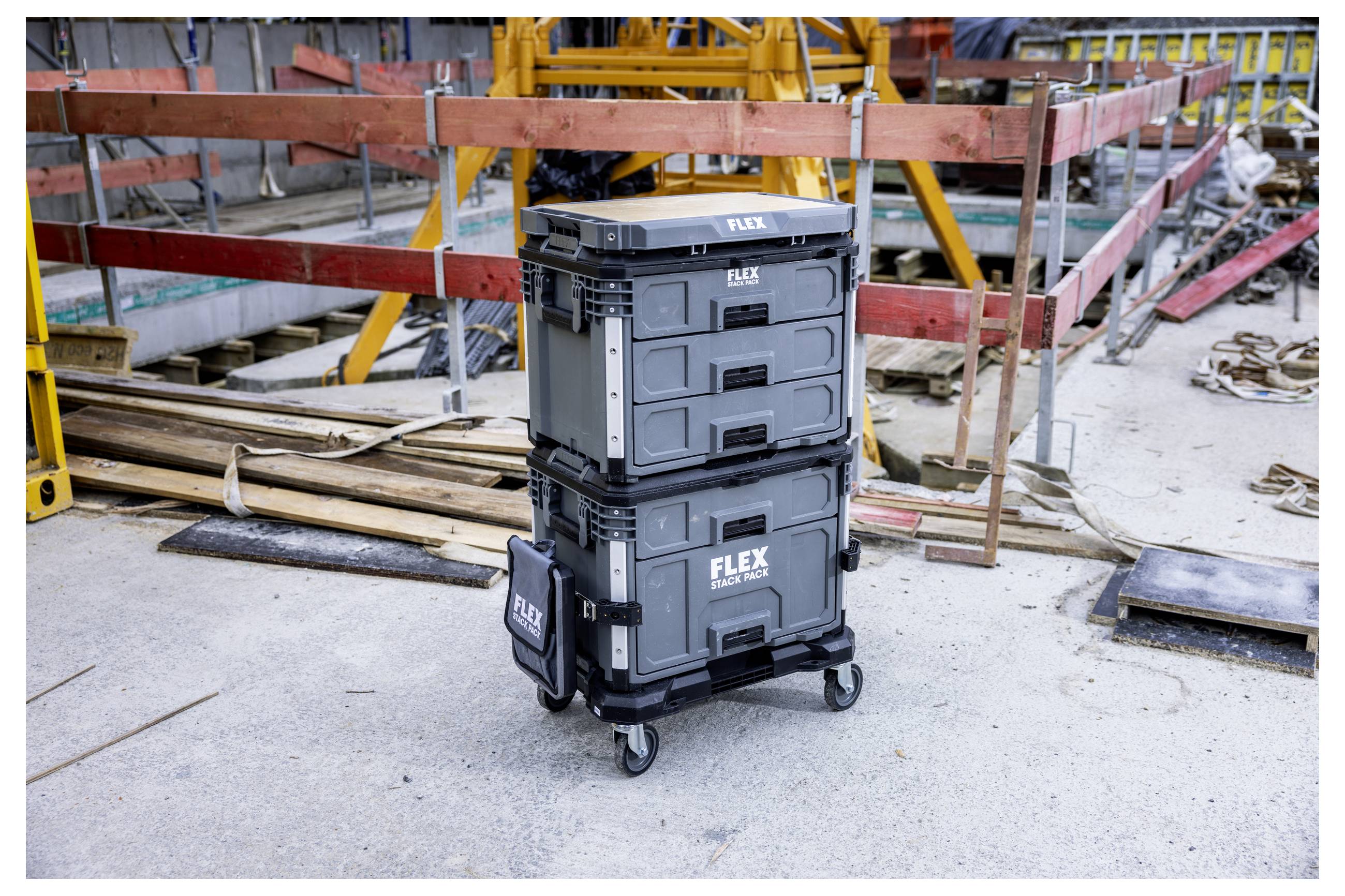 Deux caisses de rangement d'outils FLEX empilées sur un chariot à roulettes sur un chantier de construction, avec du bois de charpente, des rails et de l'équipement en arrière-plan.