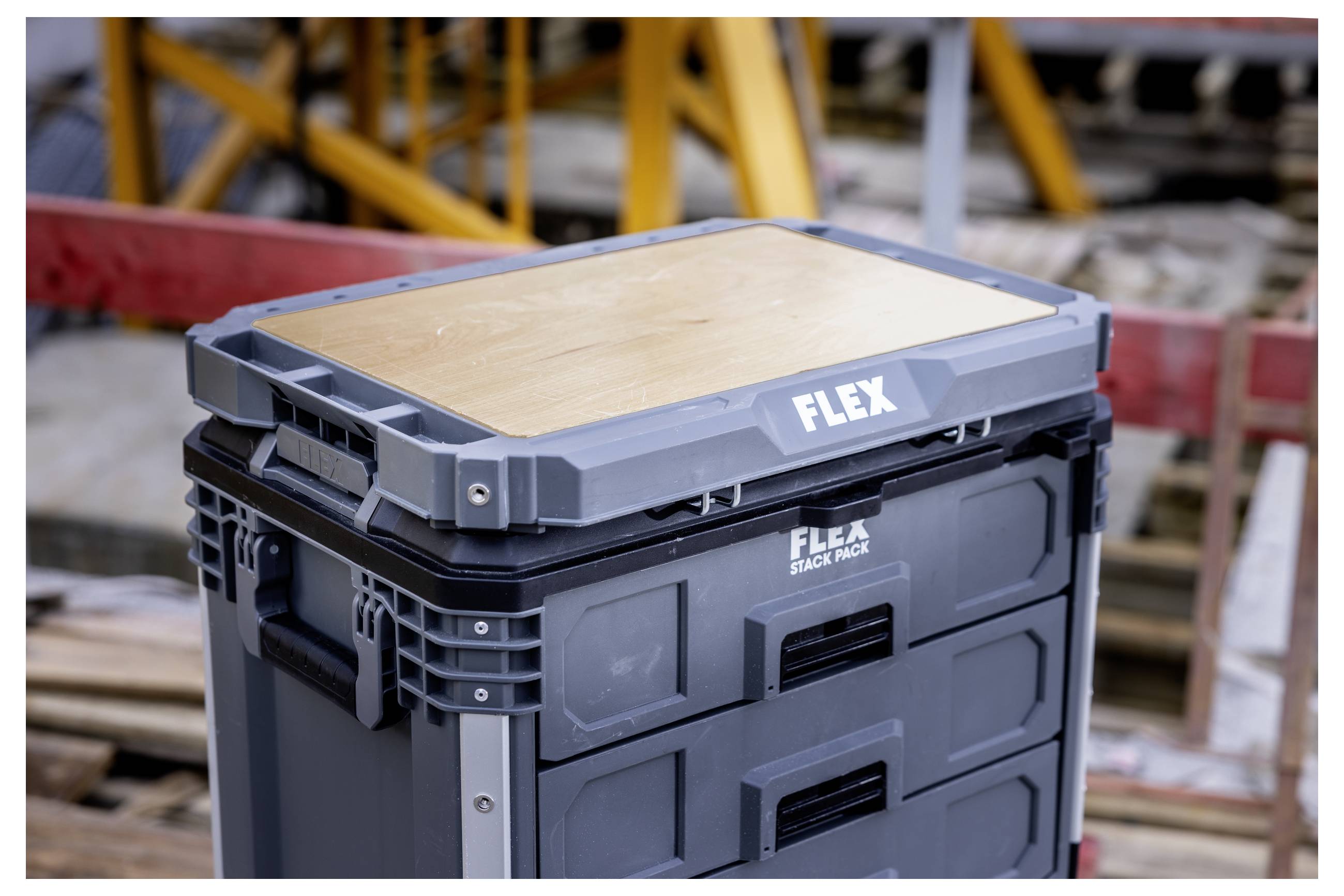 Boîtes de rangement FLEX empilées avec un dessus en contreplaqué, présentées sur un chantier avec des matériaux de construction flous en arrière-plan.