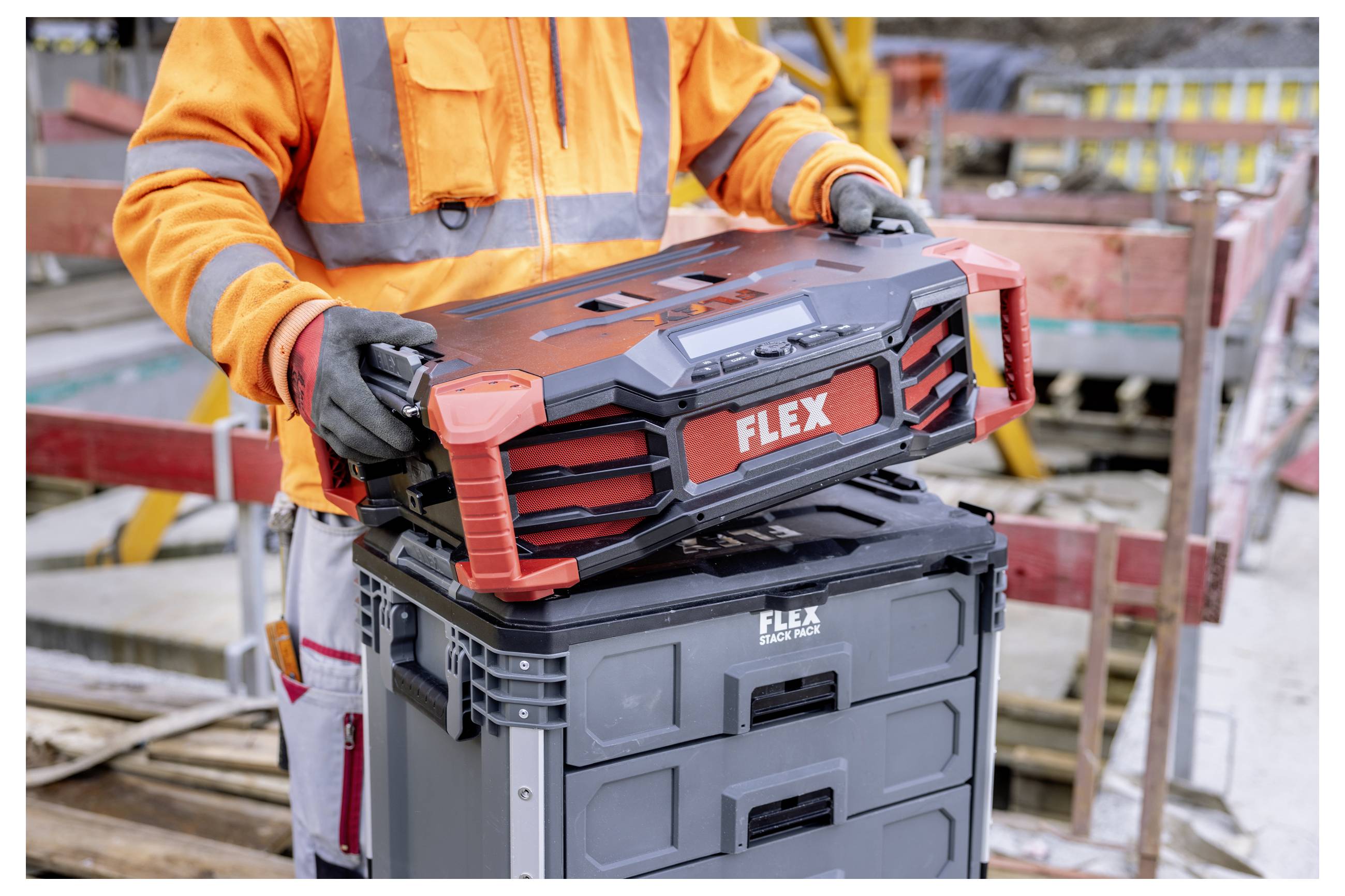 Ouvrier de la construction en équipement de sécurité orange soulevant une boîte à outils portable FLEX d'un coffre de rangement gris sur un chantier