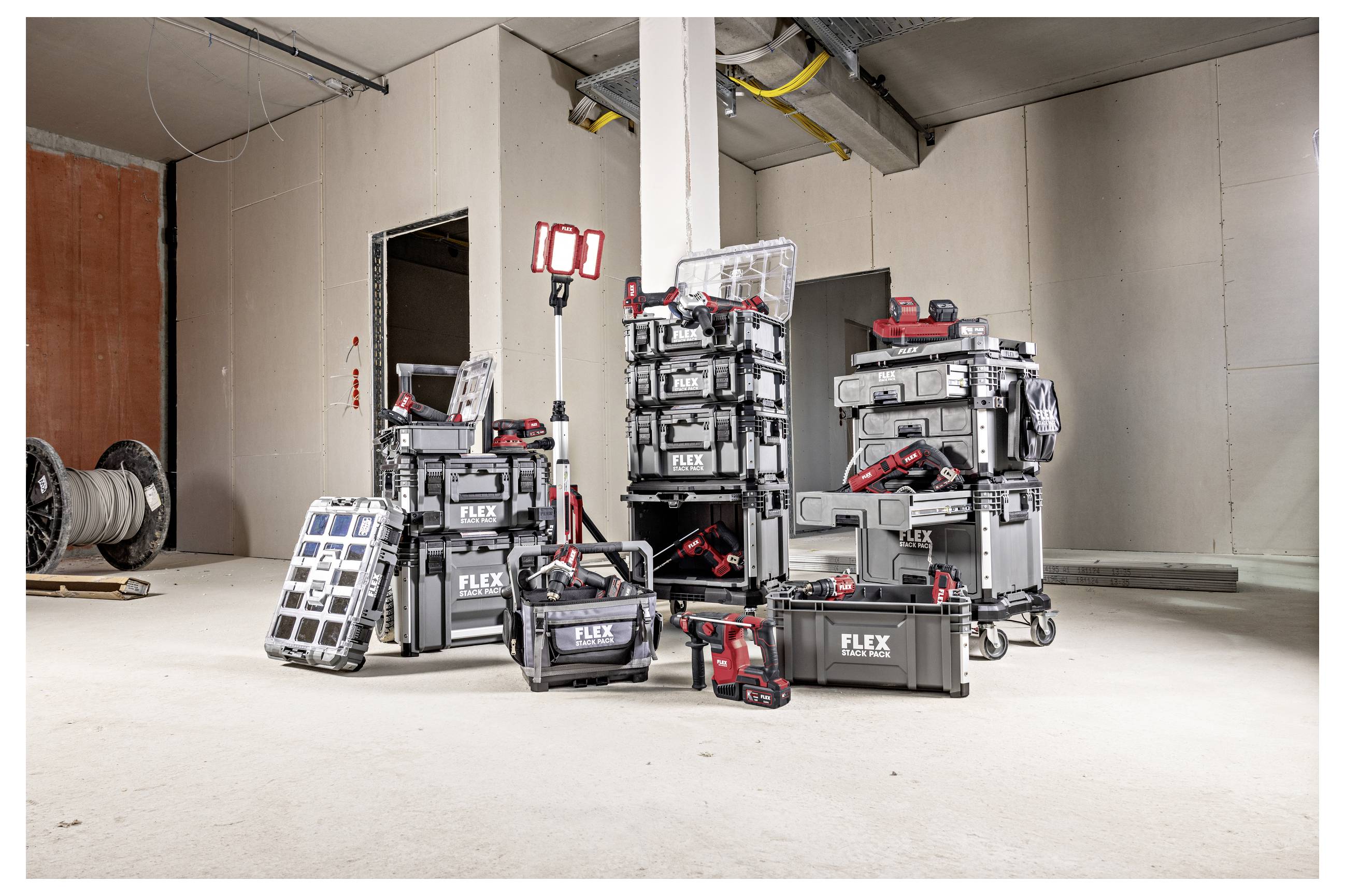Assortiment d'outils électriques FLEX et de boîtes à outils empilés dans une pièce de type chantier de construction, avec une lampe de travail et des outils à batterie exposés à l'avant.