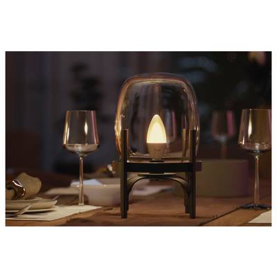 Une lampe ressemblant à une bougie scintillante à l'intérieur d'une cloche en verre repose sur une table de restaurant avec des verres de vin et des couverts dans un restaurant faiblement éclairé.