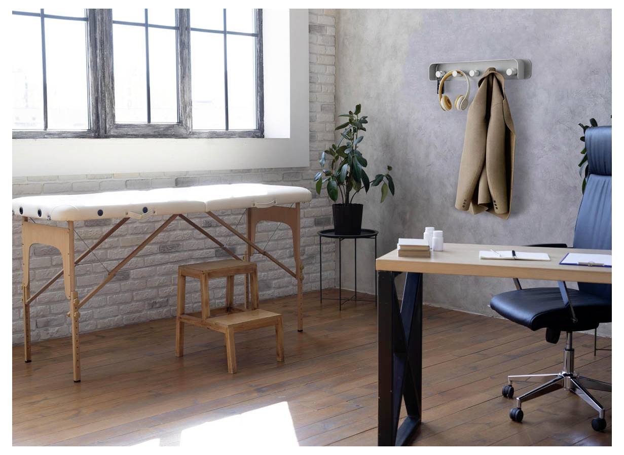 Bureau de thérapie minimaliste avec table de massage, tabouret, bureau, chaise, porte-manteau et plante dans une pièce lumineuse avec de grandes fenêtres.