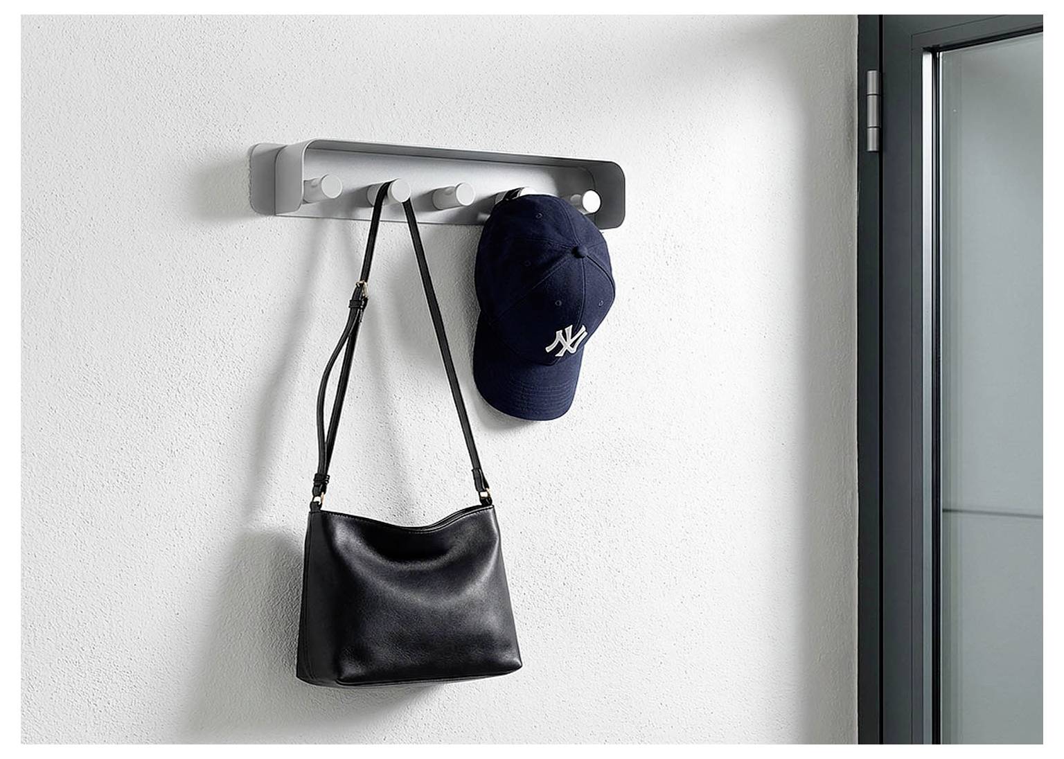 Porte-manteau mural avec un sac à main noir et une casquette de baseball marine accrochés sur un mur blanc près d'une porte.