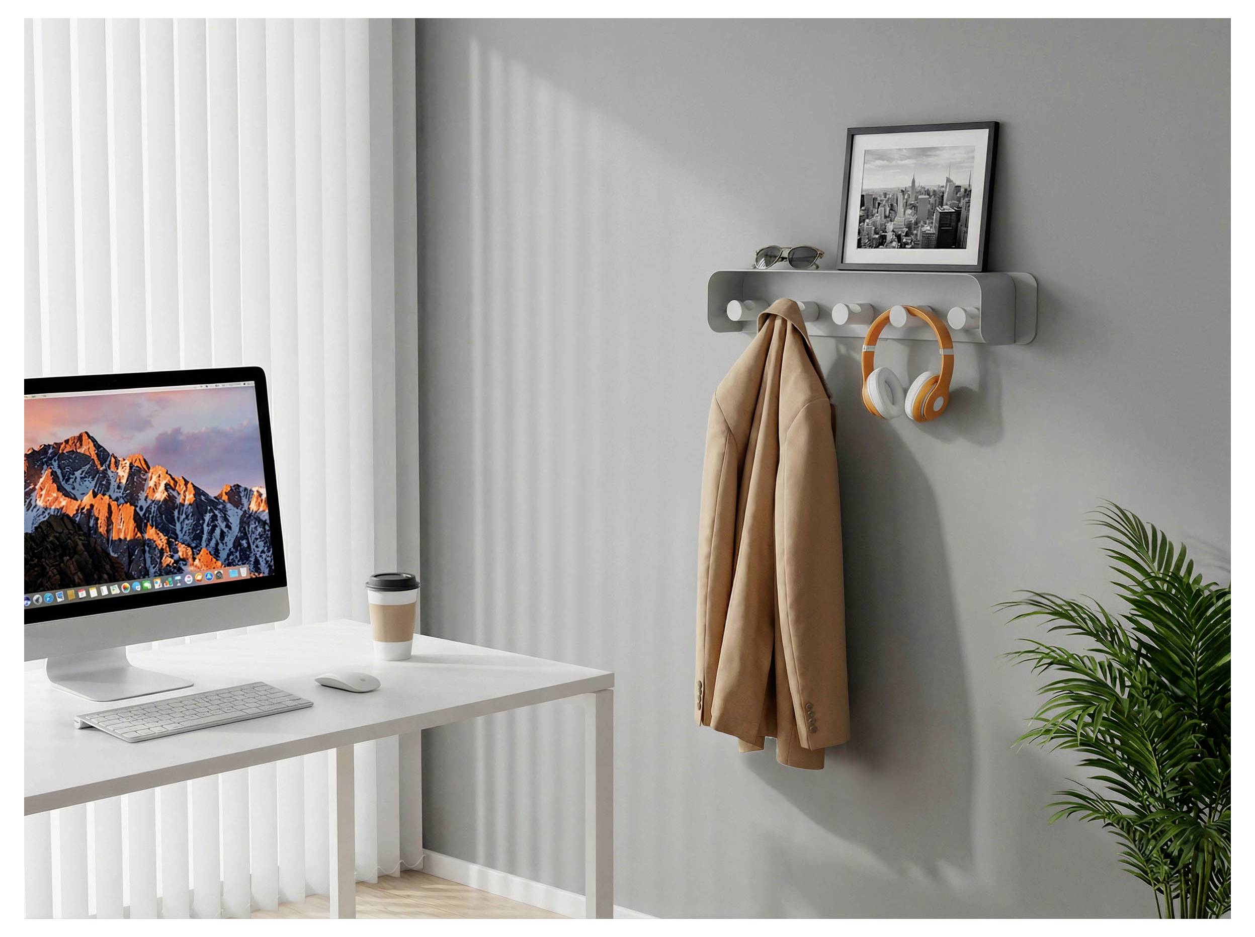 Bureau à domicile minimaliste avec un bureau, un ordinateur de bureau, une tasse de café, des crochets muraux tenant un manteau et un casque audio, une photo encadrée et une plante en pot.
