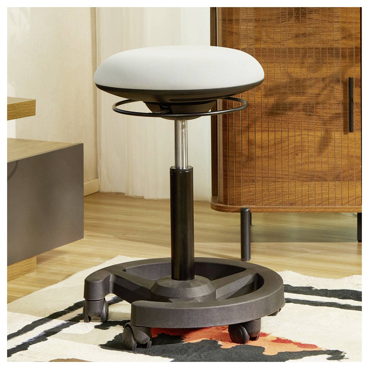 Tabouret pivotant ajustable avec siège blanc rembourré, base noire et roulettes sur un tapis à motifs dans une pièce moderne.