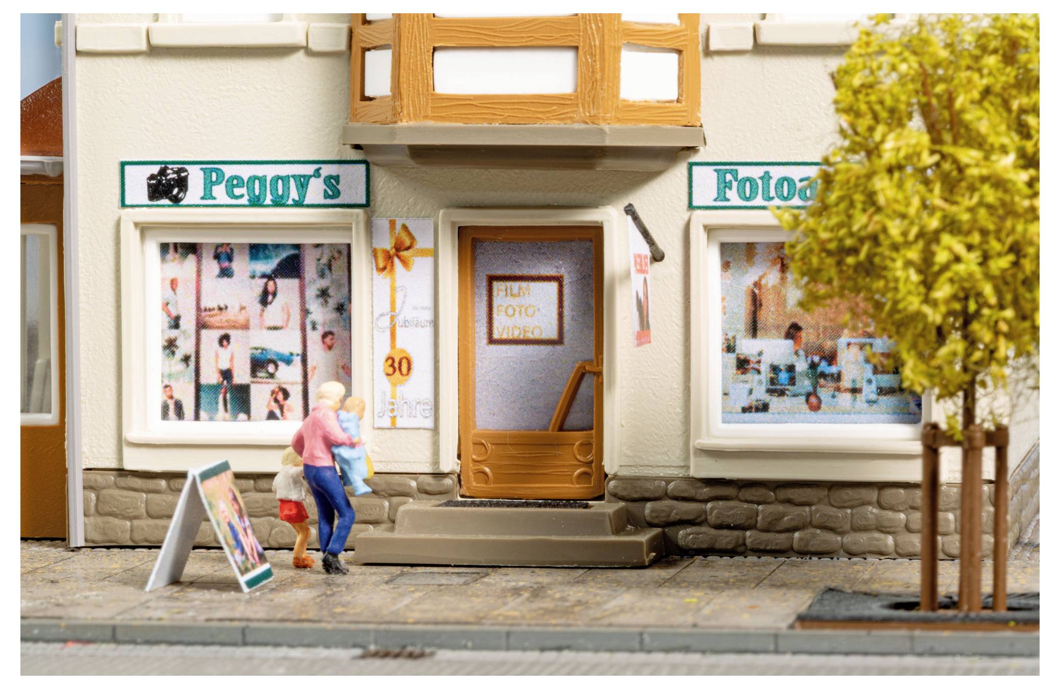 Scène de rue miniature d'un magasin de photographie appelé « Peggy's Foto » avec deux piétons marchant devant l'entrée et les vitrines d'exposition.