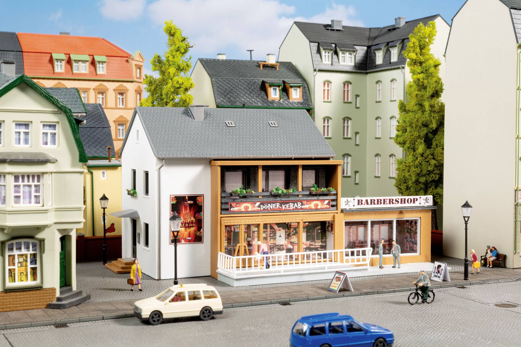 Scène de rue miniature avec des magasins, notamment « Döner Kebab » et « BARBERSHOP », un cycliste, des piétons et des voitures garées sur une route tranquille.