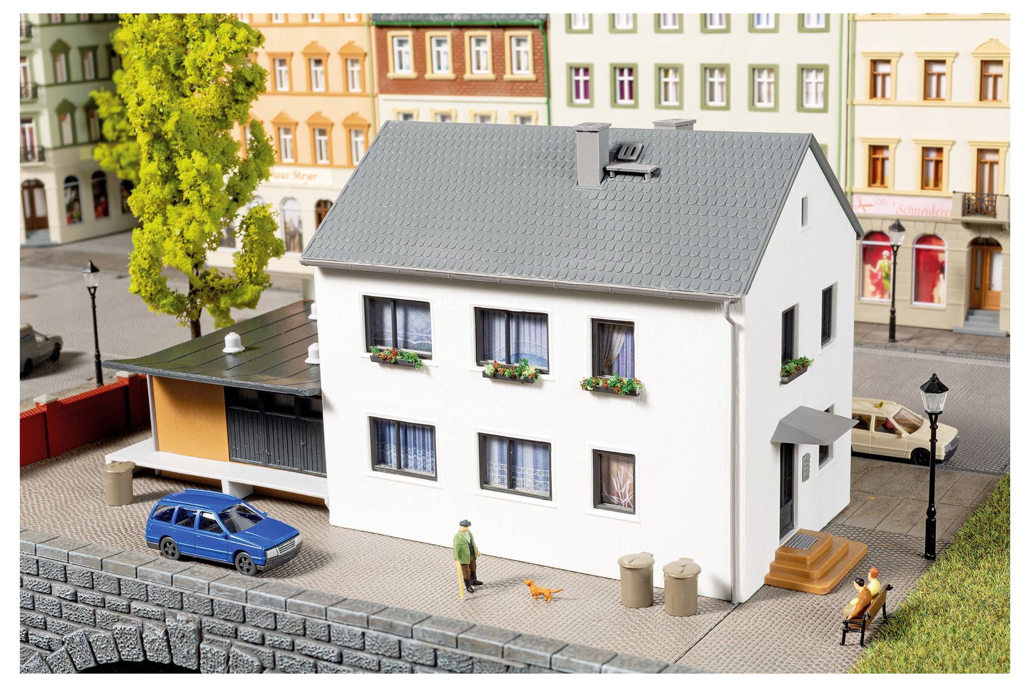Scène de ville modèle avec une maison blanche à deux étages à côté d'une route, une voiture bleue, des piétons, des bancs, des arbres et des immeubles d'appartements colorés en arrière-plan.