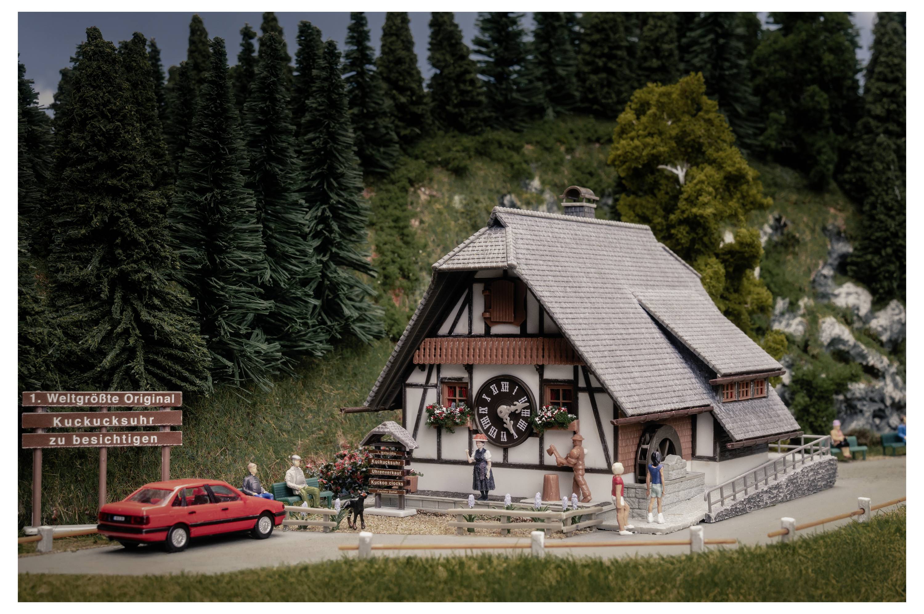 Attraction miniature de maison de montagne avec des visiteurs, une voiture rouge, et un panneau indiquant « Weltgroesste Original Kuckucksuhr zu besichtigen » à côté d'une façade géante d'horloge à coucou.