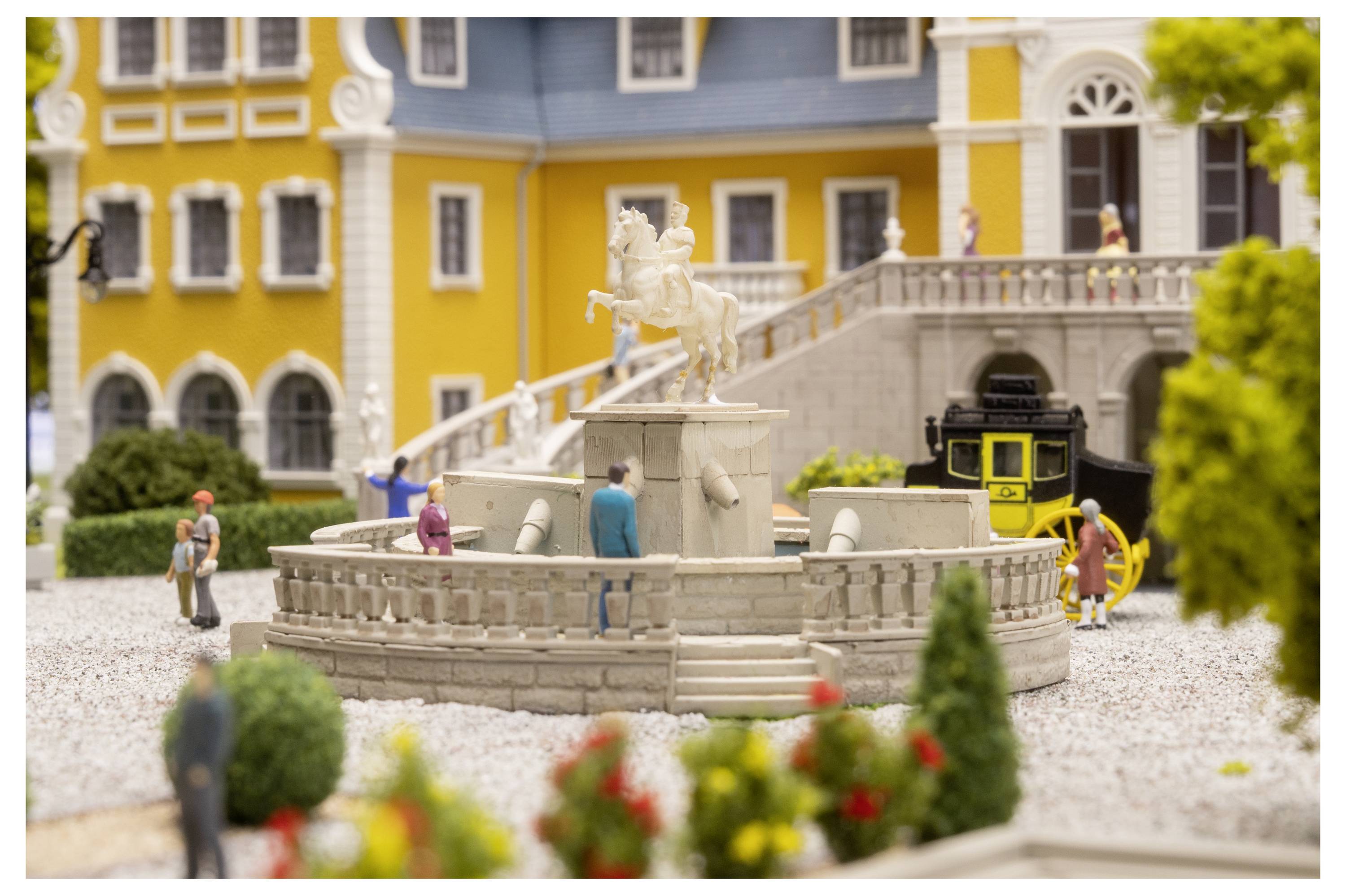 Scène extérieure miniature avec une fontaine en pierre centrale et une statue, de minuscules personnes autour, et un tramway jaune à côté d'un bâtiment jaune et d'un escalier gris.