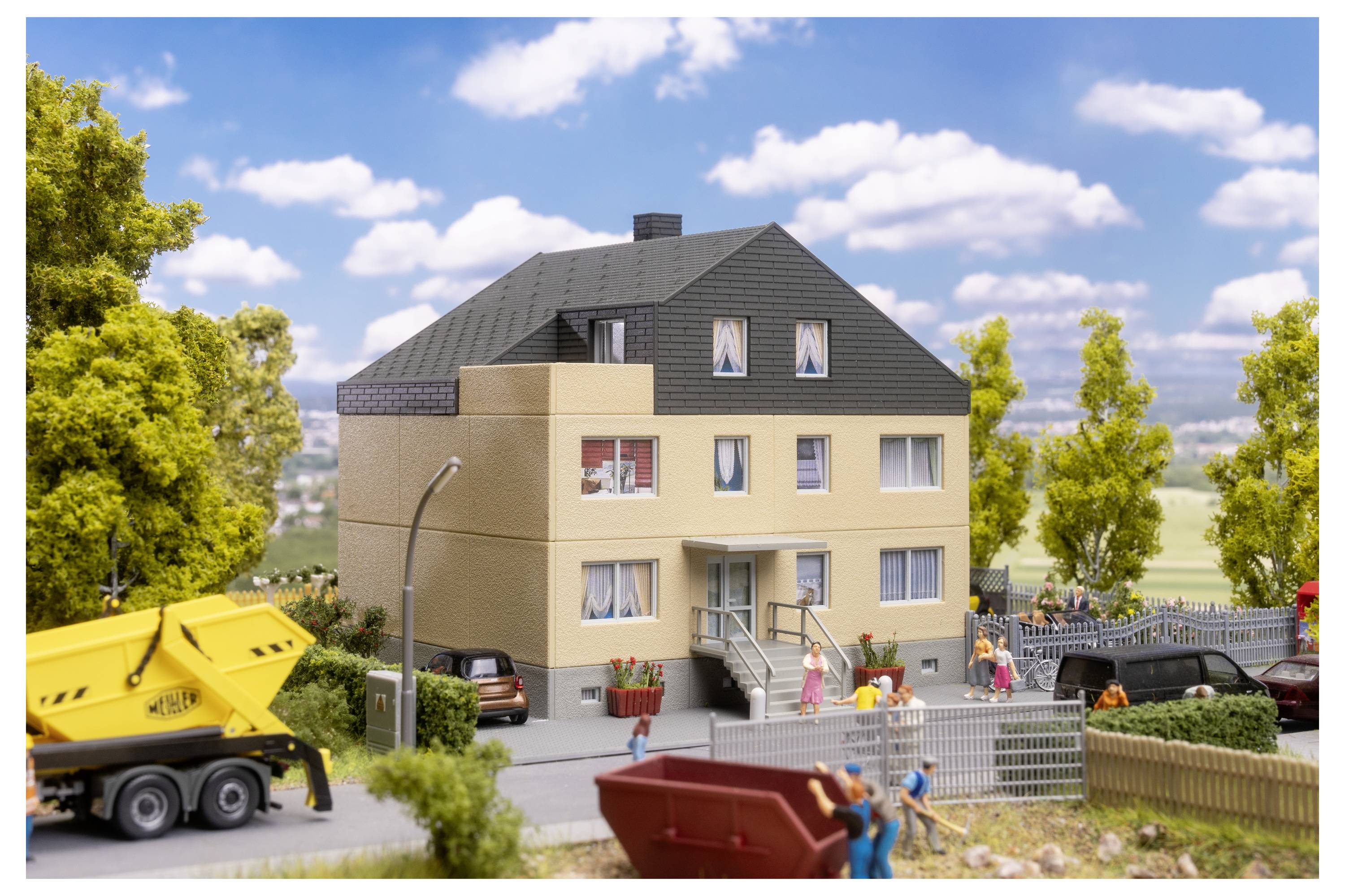 Scène résidentielle miniature avec un immeuble d'appartements beige, des voitures garées, des ouvriers et un camion à benne jaune sous un ciel bleu.