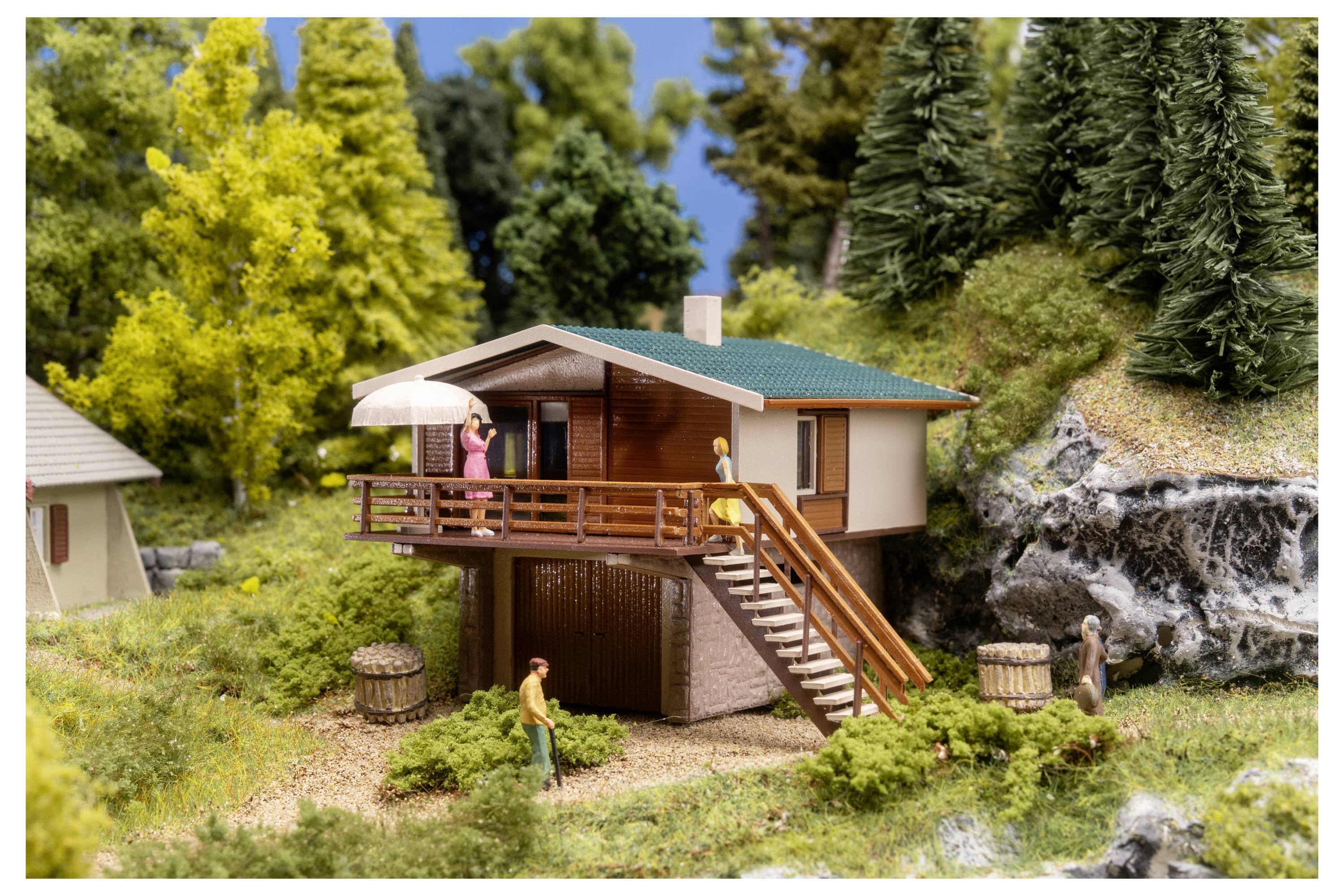 Maquette miniature d'une maison sur un versant avec des escaliers extérieurs, de minuscules figurines et des arbres et rochers environnants.