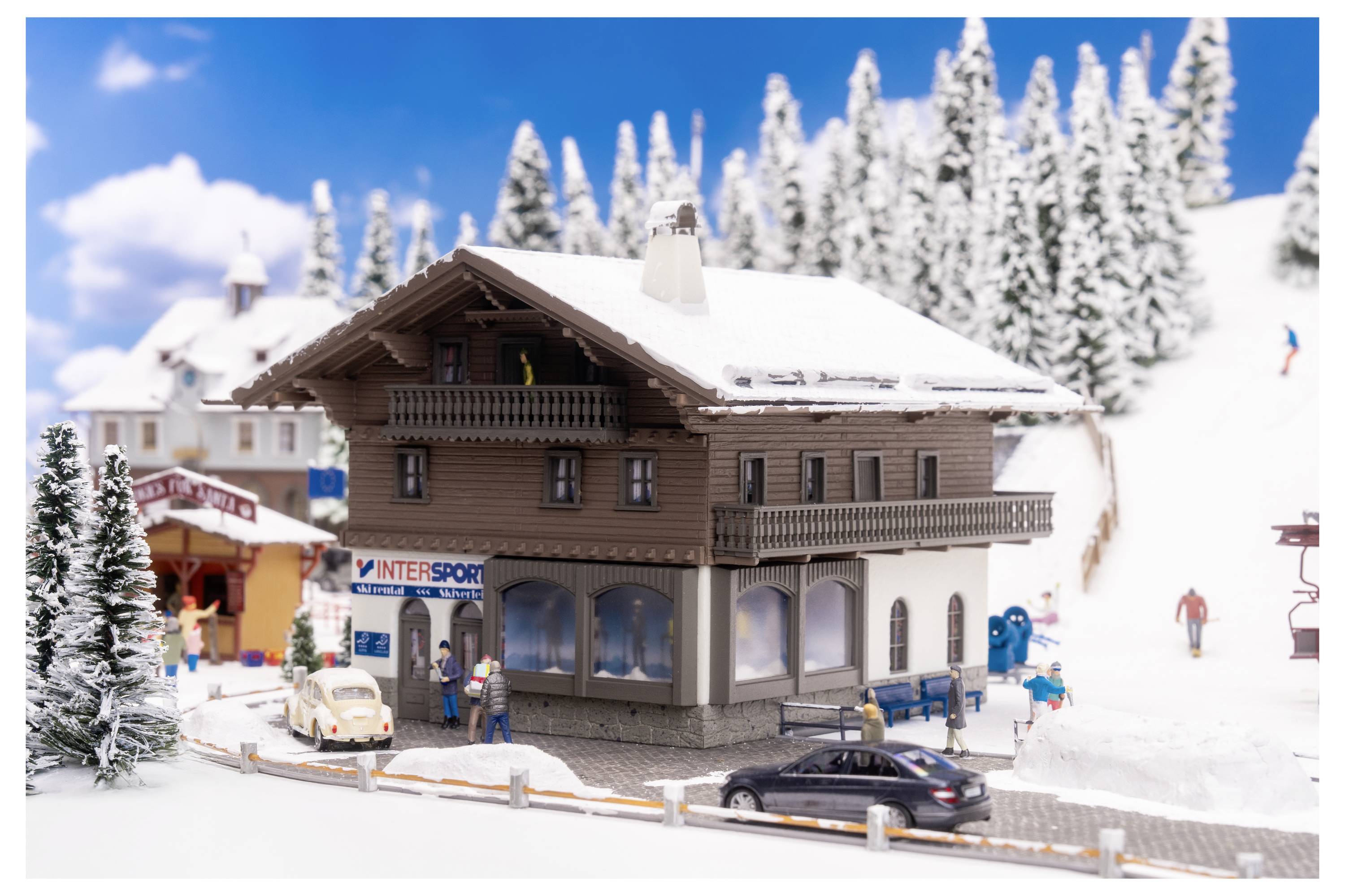Un petit village de station de ski enneigée avec des bâtiments de chalets, des voitures garées et des skieurs sur un versant montagneux hivernal ensoleillé.