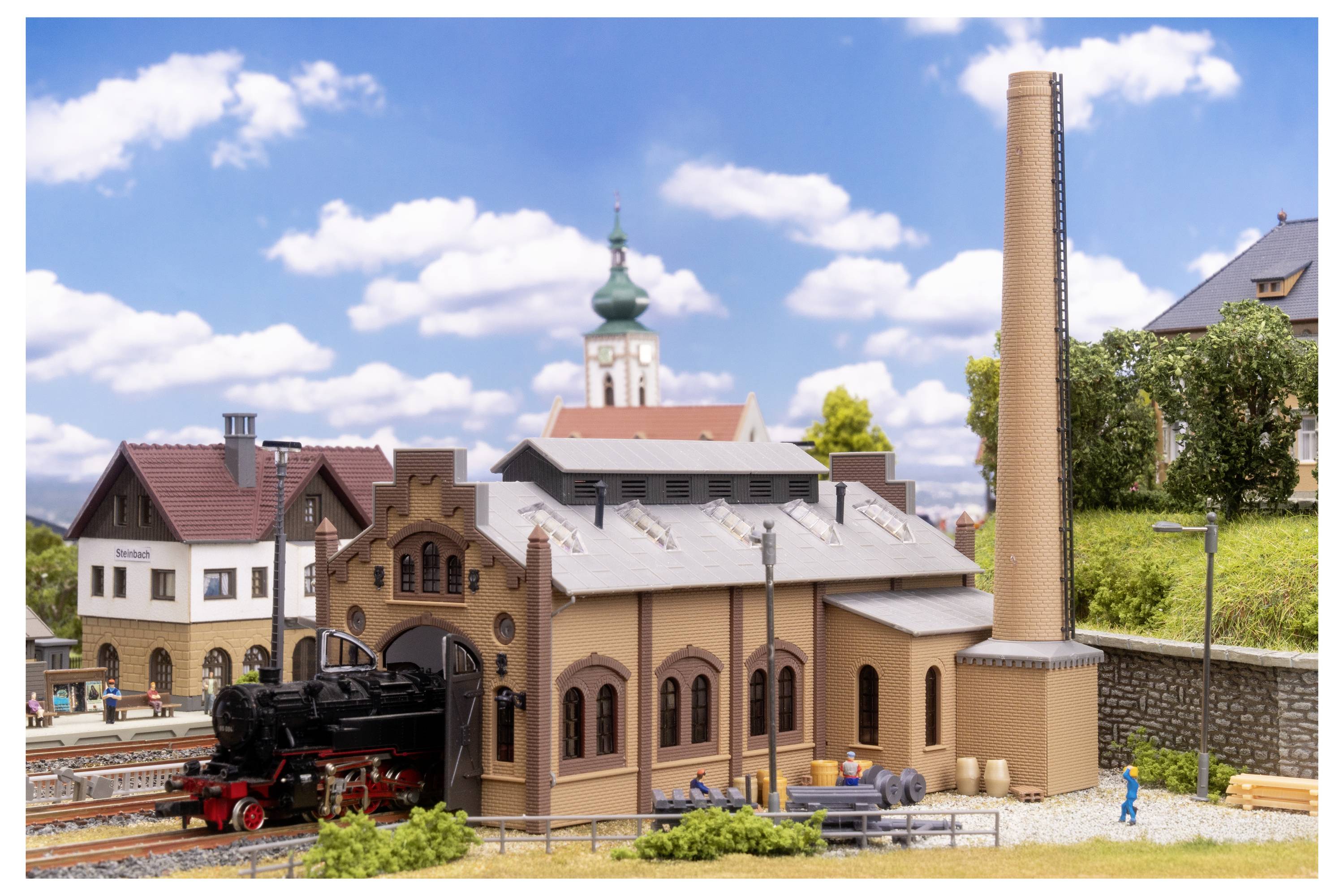 Scène de chemin de fer miniature avec une locomotive à vapeur à côté d'un dépôt en forme d'arche, une haute cheminée, et des bâtiments villageois sous un ciel bleu.