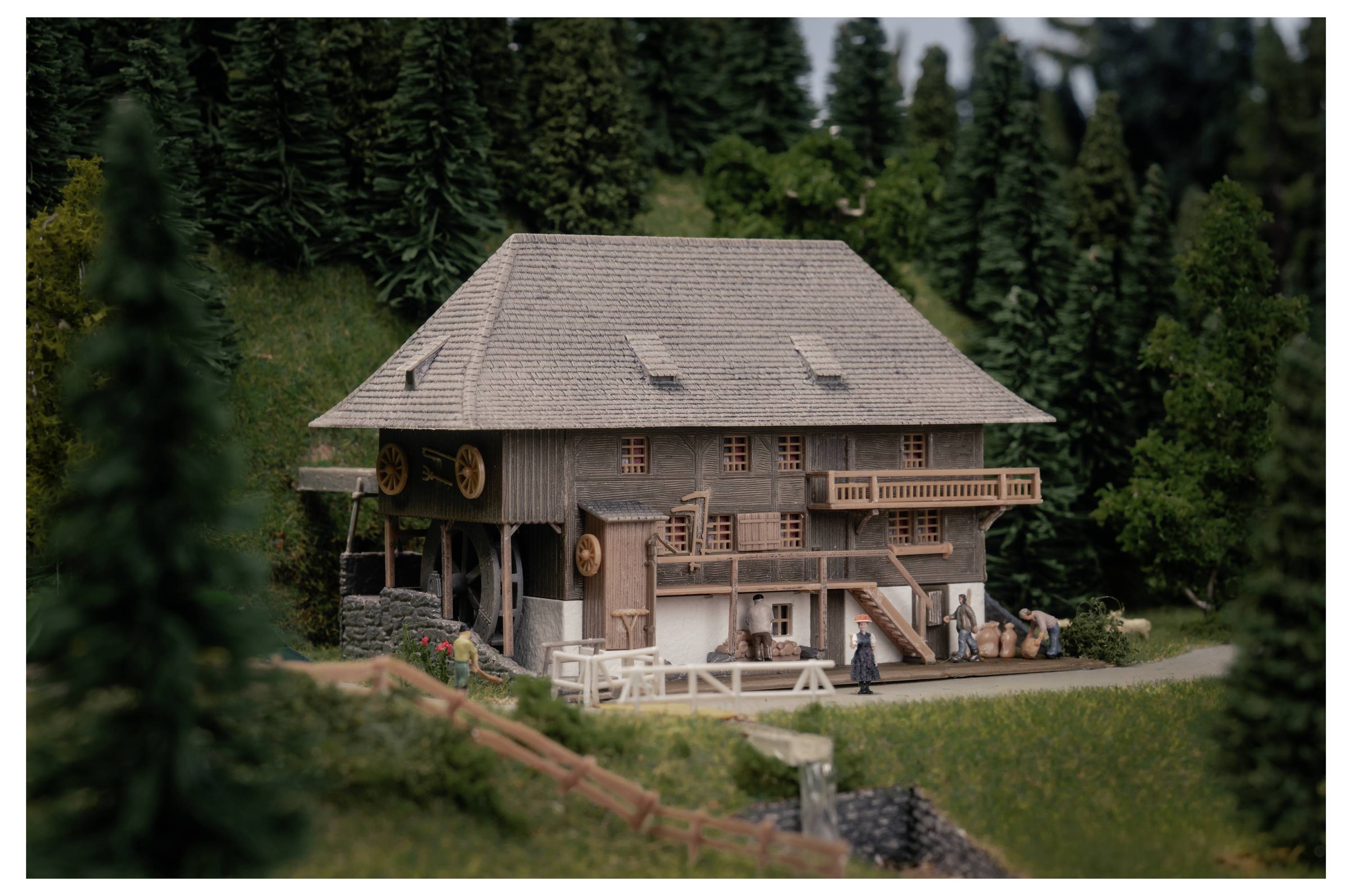 Un modèle miniature d'une maison en bois rustique avec des balcons et un toit en pente, situé au milieu de denses sapins persistants et d'un paysage herbeux.