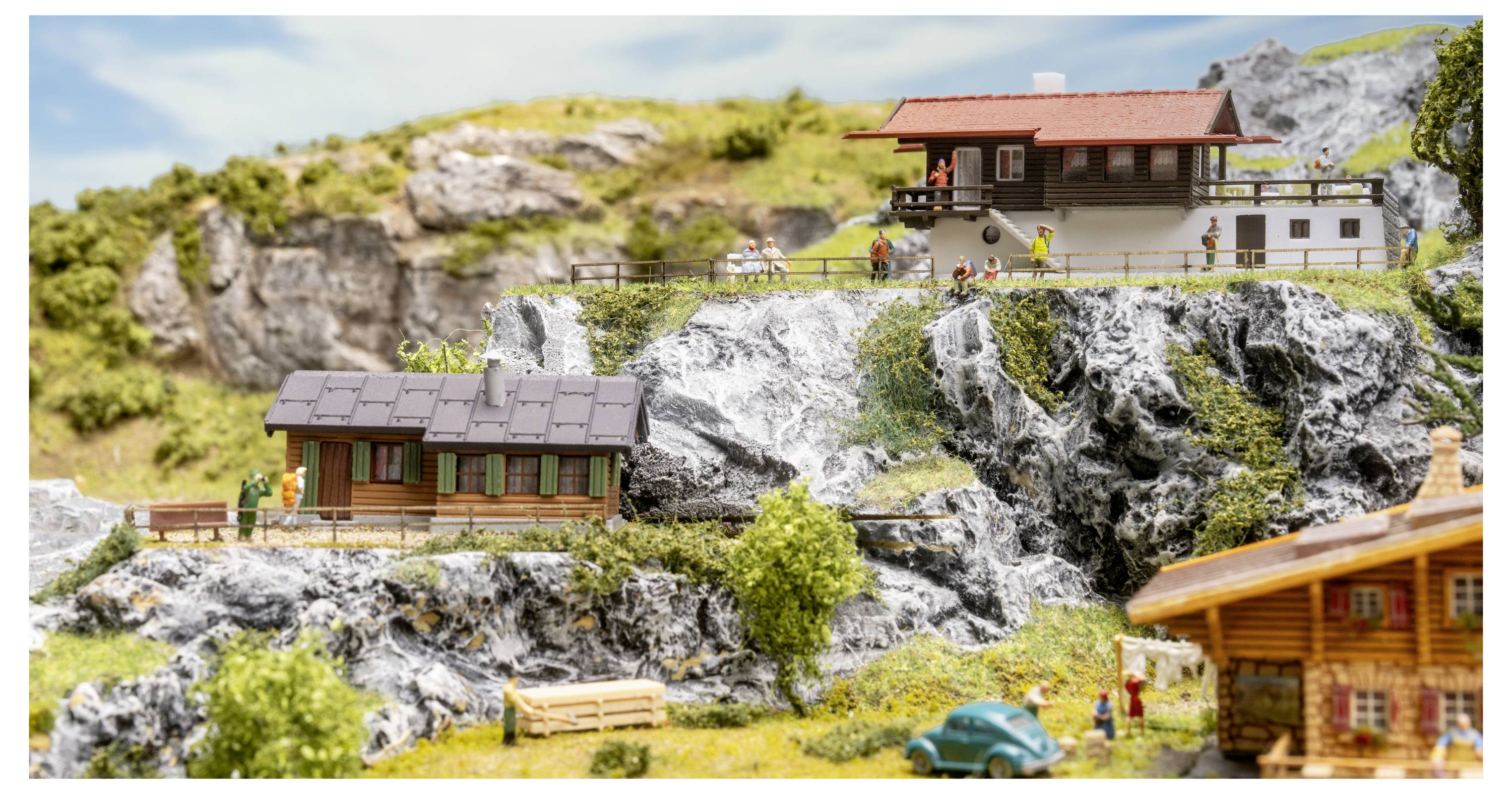 Maquette de village montagnard miniature avec chalets en bois sur des pentes rocheuses, une maison en bord de falaise, de minuscules figurines, des arbres et une petite voiture bleue.