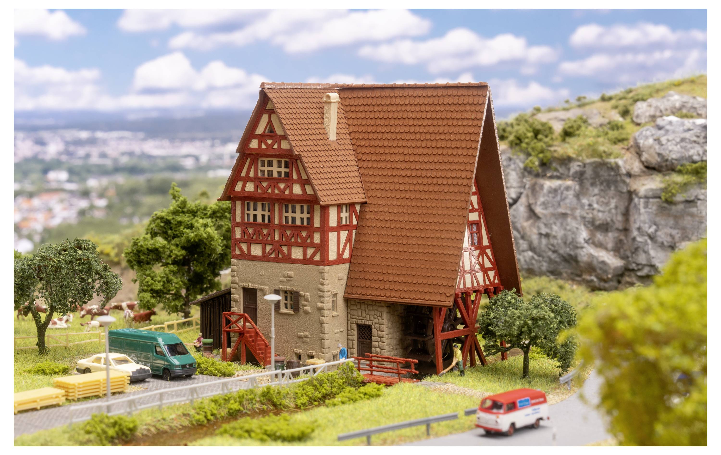 Modèle détaillé d'une maison à colombages avec toit rouge sur un paysage miniature rural, avec des arbres, une route et de petits véhicules à proximité.