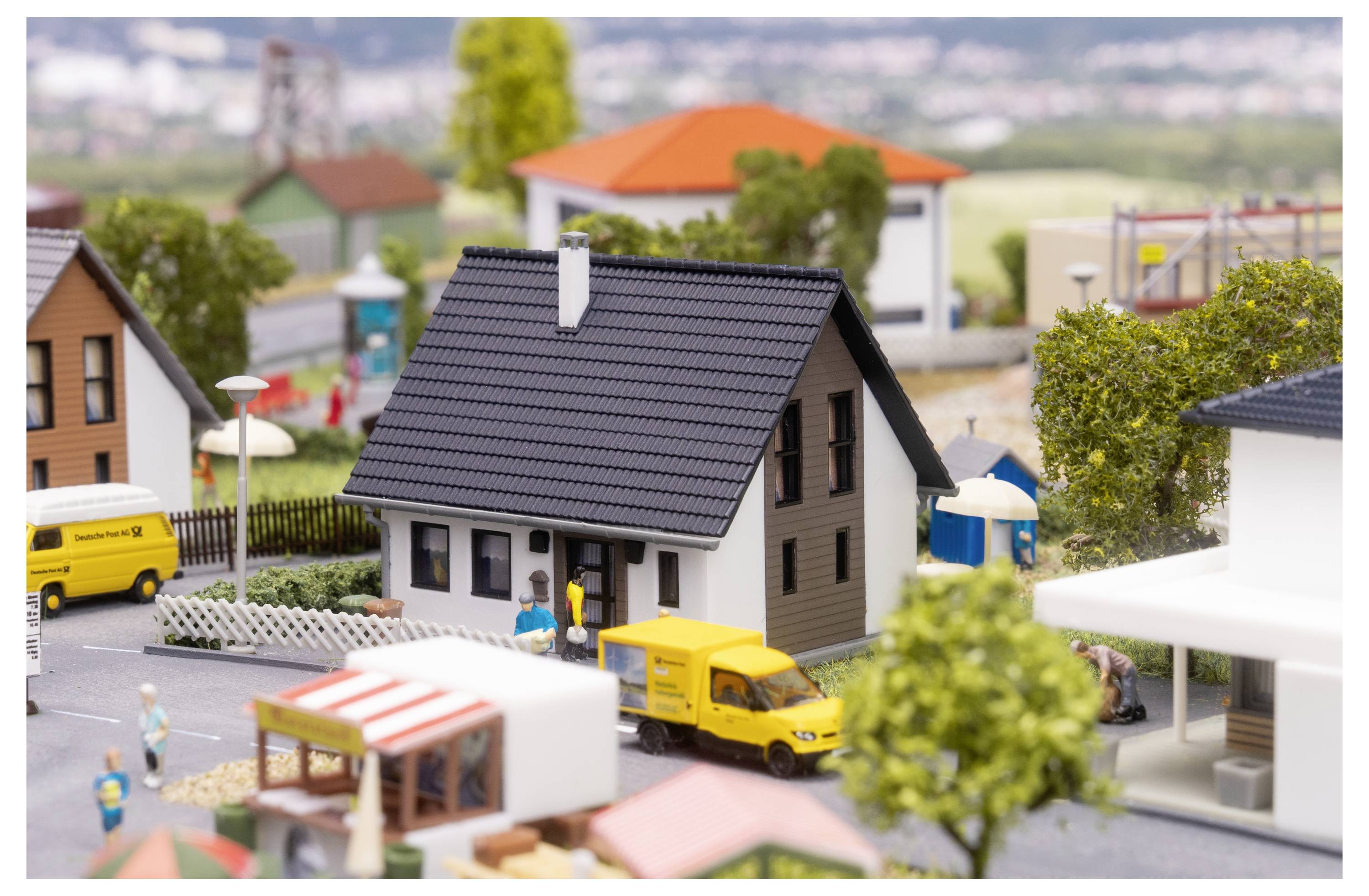 Maquette miniature d'un quartier résidentiel avec une maison grise centrale, un fourgon de livraison jaune et de petites figurines dans la rue.