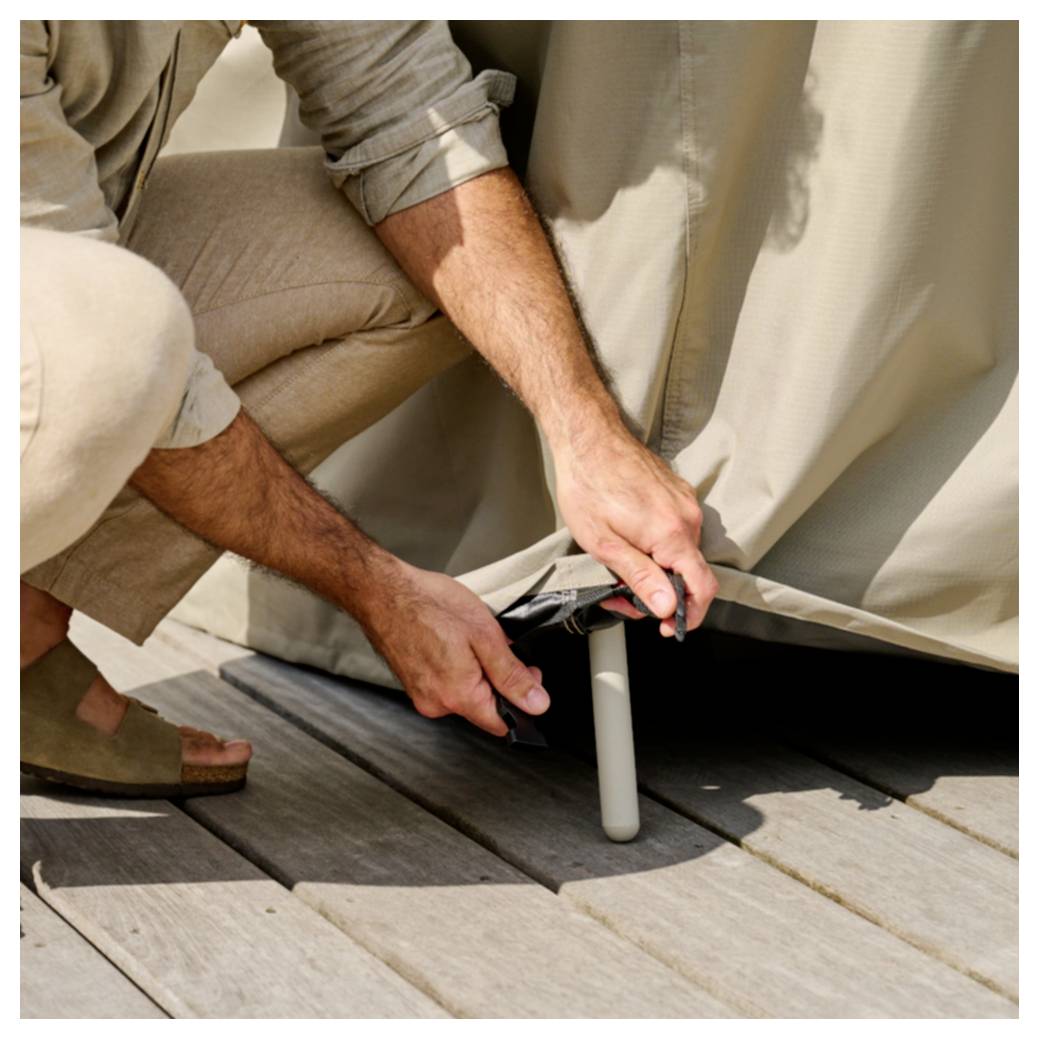 Une personne s'accroupit pour attacher ou fixer une housse de mobilier d'extérieur beige autour d'une jambe de chaise sur une terrasse en bois.