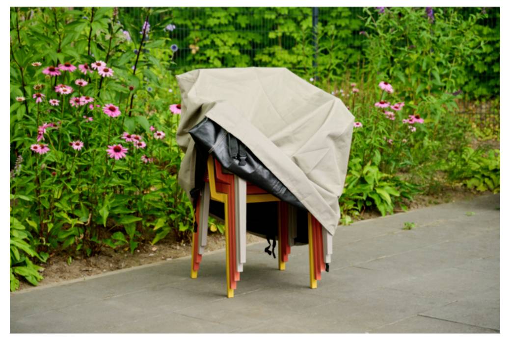Des chaises empilées recouvertes d'une bâche beige à l'extérieur, à côté d'un chemin pavé et d'un jardin fleuri.