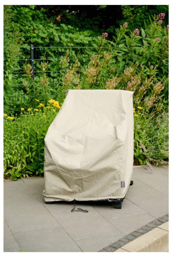 Une housse de protection beige recouvre une chaise d'extérieur ou un grill à roulettes sur un patio, avec des plantes de jardin en arrière-plan.