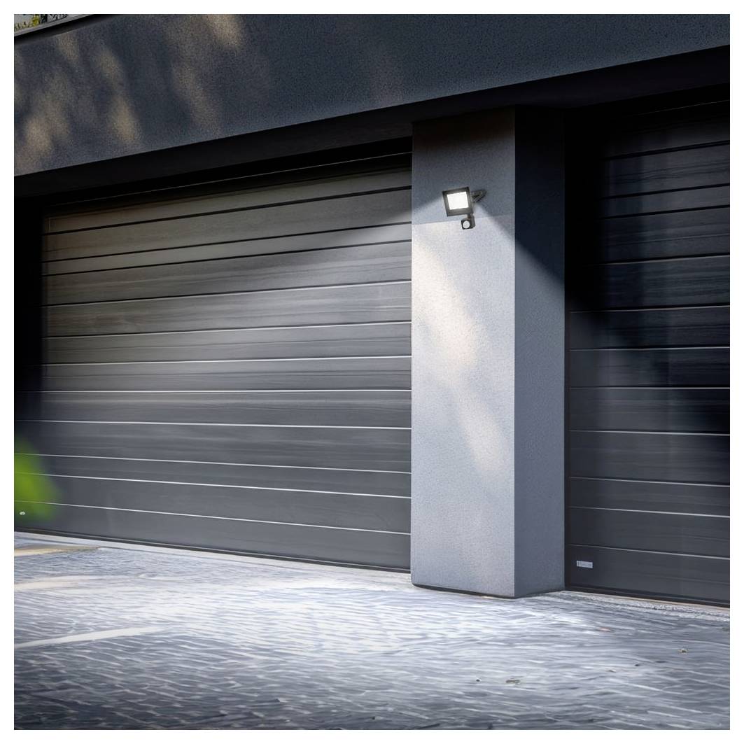 Extérieur de maison moderne avec deux portes de garage fermées de couleur foncée et une lampe murale illuminée à côté du pilier d'entrée.