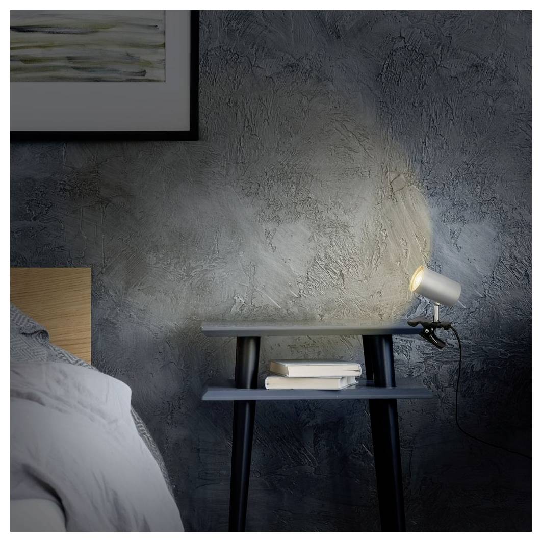 Table de nuit dans une chambre à coucher avec une lampe projecteur allumée, des livres empilés, et une partie d'un lit contre un mur texturé sombre.