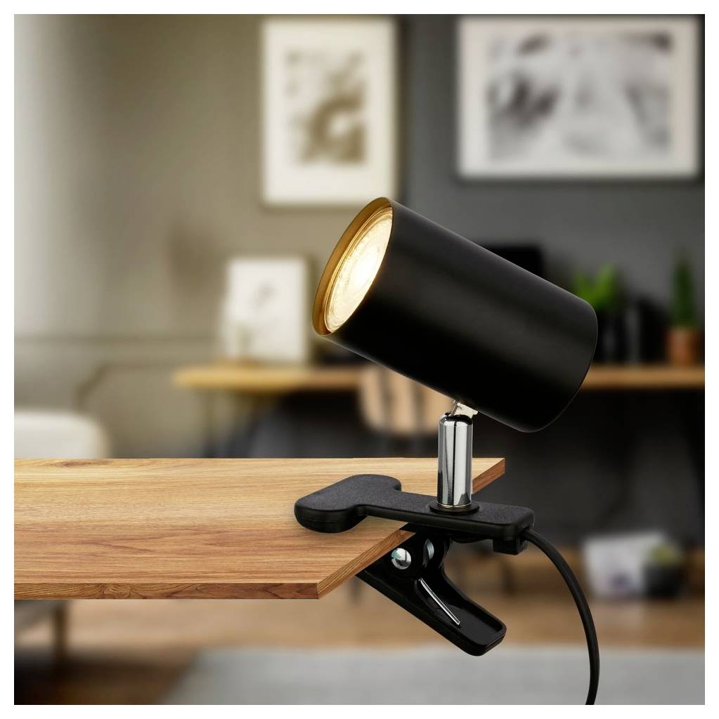 Lampe projecteur noir à pince fixée au bord d'un bureau en bois, orientée pour éclairer l'espace de travail.