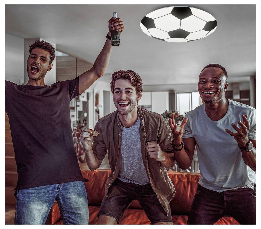 Trois hommes applaudissent sur un canapé, l'un d'eux tenant une bouteille, tandis qu'ils regardent un match de football à la télévision dans un salon.