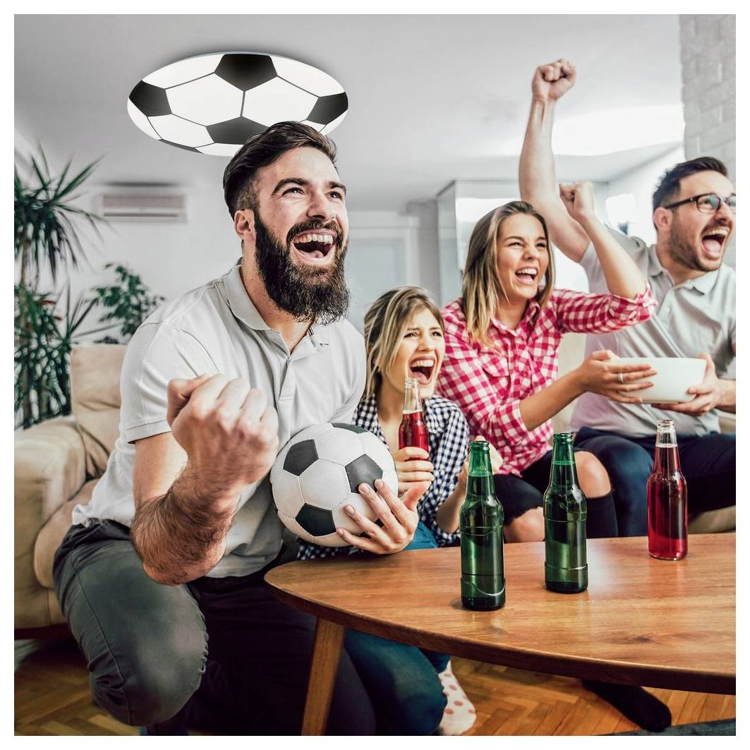 Des amis qui crient de joie à la maison en regardant un match de soccer, un homme tenant un ballon et d'autres qui célèbrent sur un canapé avec des boissons à proximité.