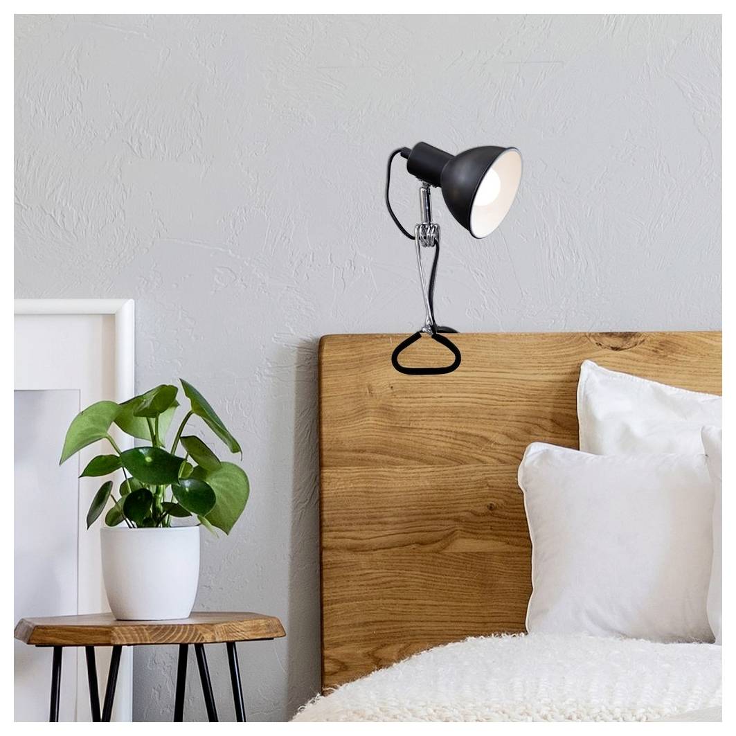 Coin de chambre minimaliste avec tête de lit en bois, lampe murale noire allumée, oreillers blancs, plante en pot et tableau encadré appuyé contre le mur.
