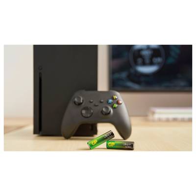 Console Xbox noire avec une manette et deux piles GP Ultra AA sur un bureau devant un écran.