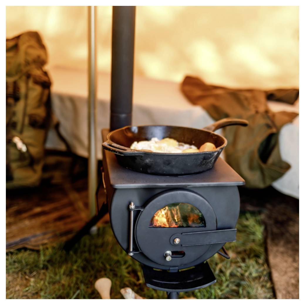 Un petit poêle de camping fonctionnant au bois qui chauffe une poêle contenant de la nourriture, installé dans un confortable campement sous tente.
