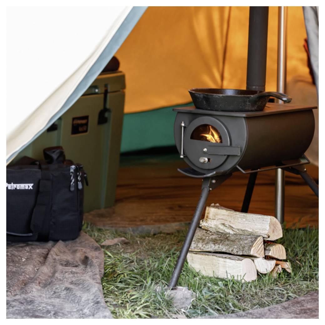 Petit poêle à bois avec une casserole sur le dessus à l'intérieur d'une tente, avec le feu visible à travers la porte et du bois de chauffage fendu empilé à côté.
