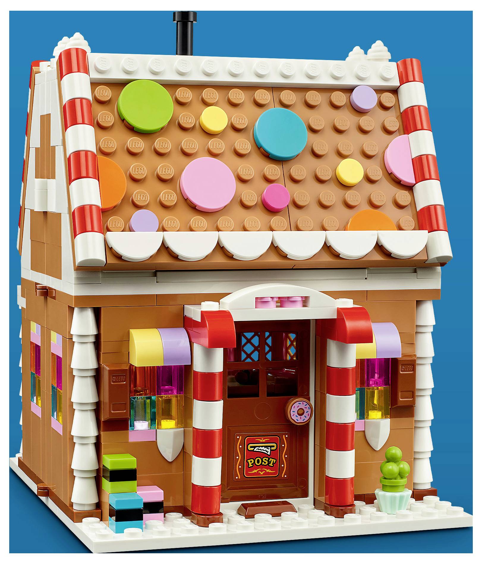 Une maison de pain d'épices colorée en LEGO avec des décorations de bonbons, des colonnes rayées, une porte d'entrée étiquetée « POSTE », et une petite figurine en pot près de l'entrée.