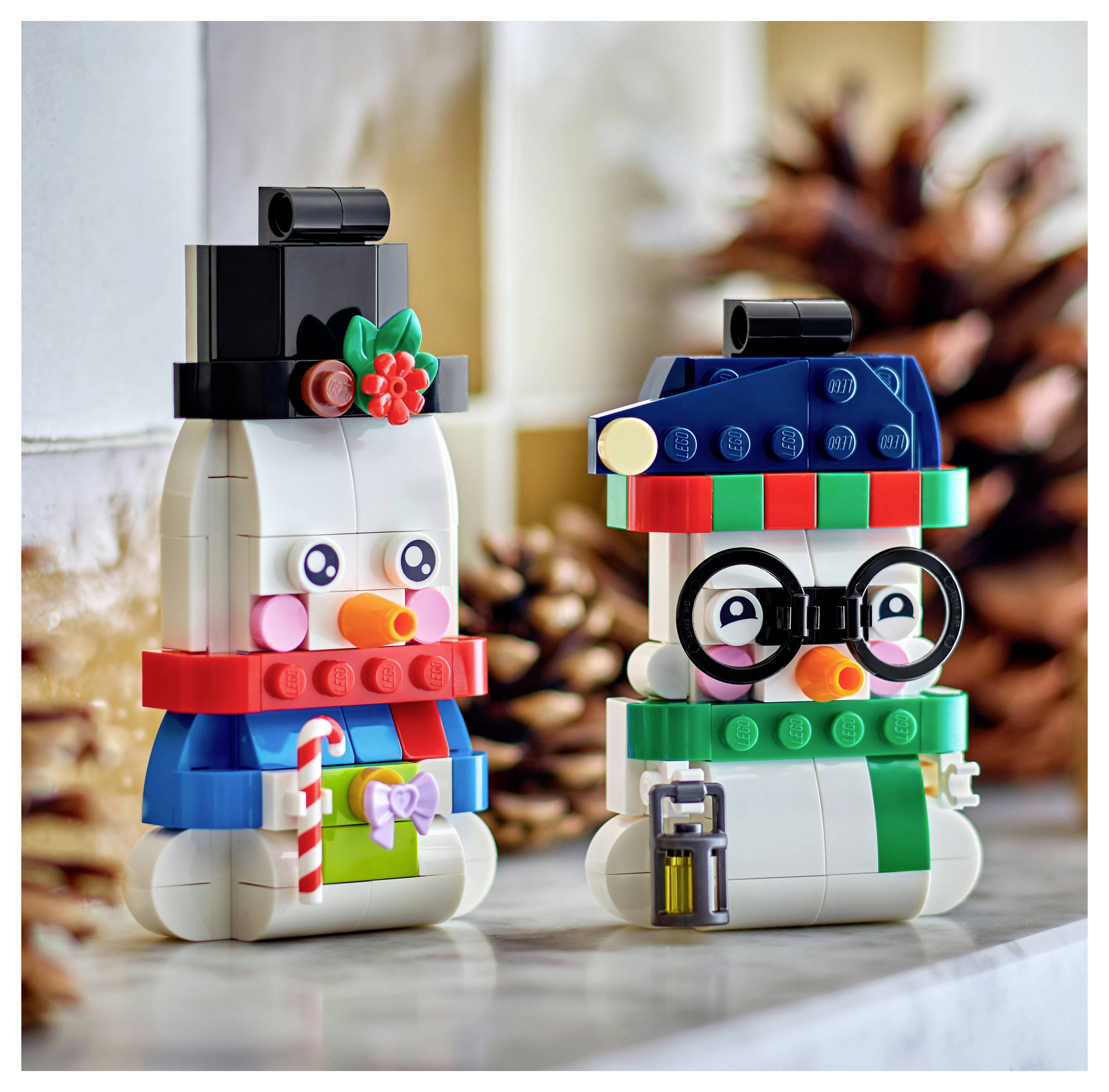 Deux figurines de bonhomme de neige LEGO portant des chapeaux, des écharpes et des lunettes sont assis sur un plateau de table, l'une tenant une canne à sucre et un cadeau, sur un fond de pomme de pin.
