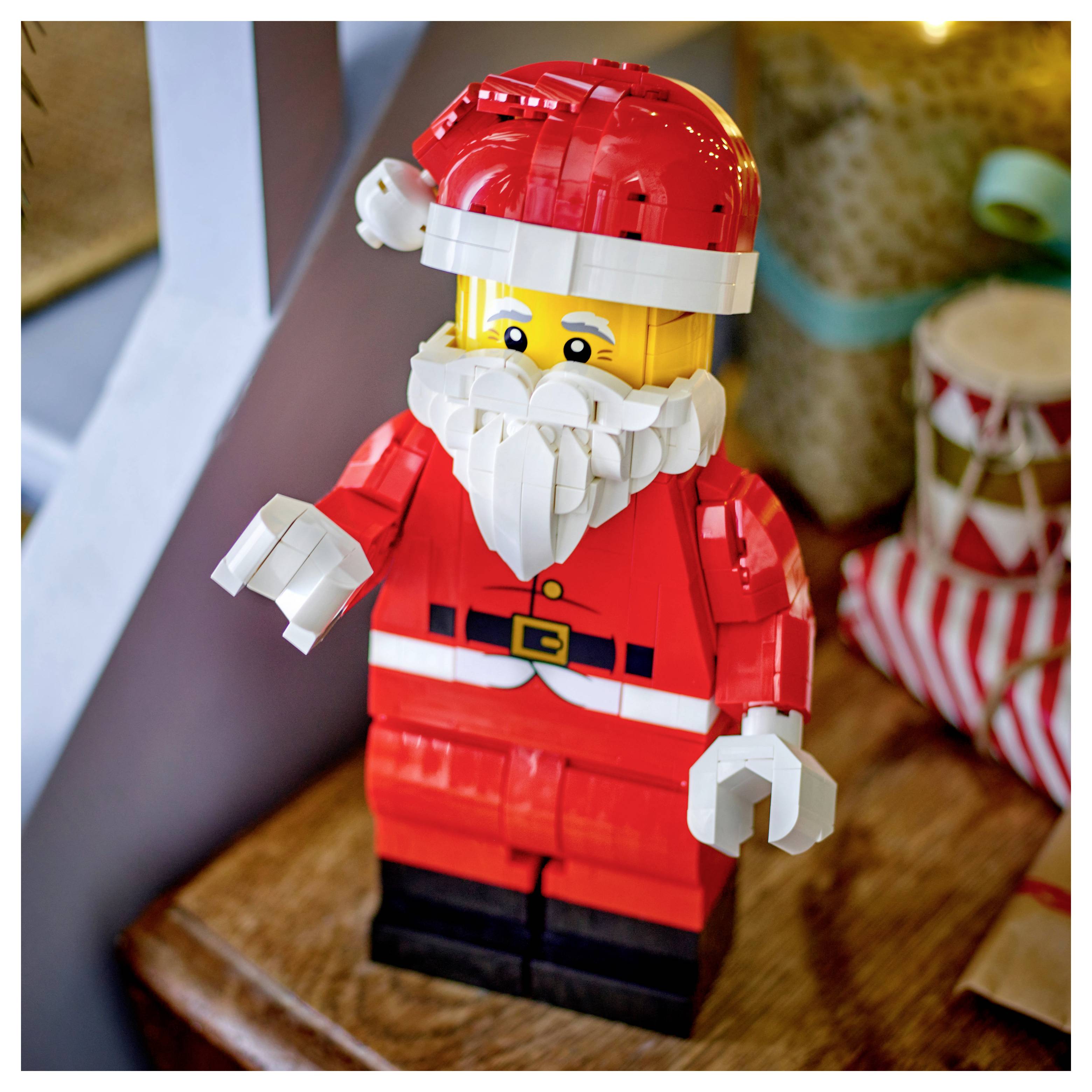 Figurine LEGO du Père Noël dans un costume et un chapeau rouges, avec une barbe blanche, se tenant debout sur une surface en bois à côté d'un cadeau emballé.