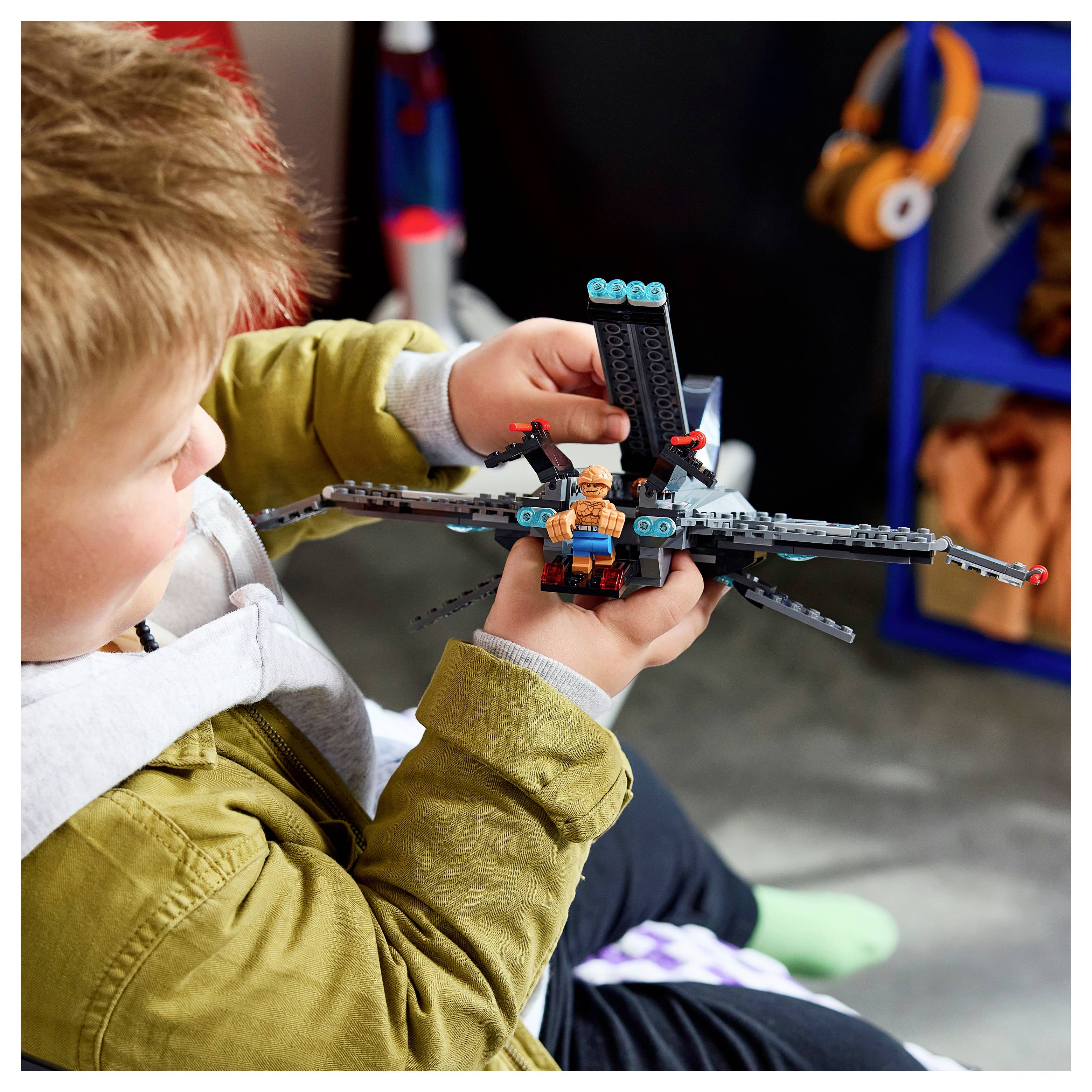 Jeune garçon tenant et inspectant un avion jouet ou un drone de style LEGO avec une figurine miniature, assis à l'intérieur en train de jouer.