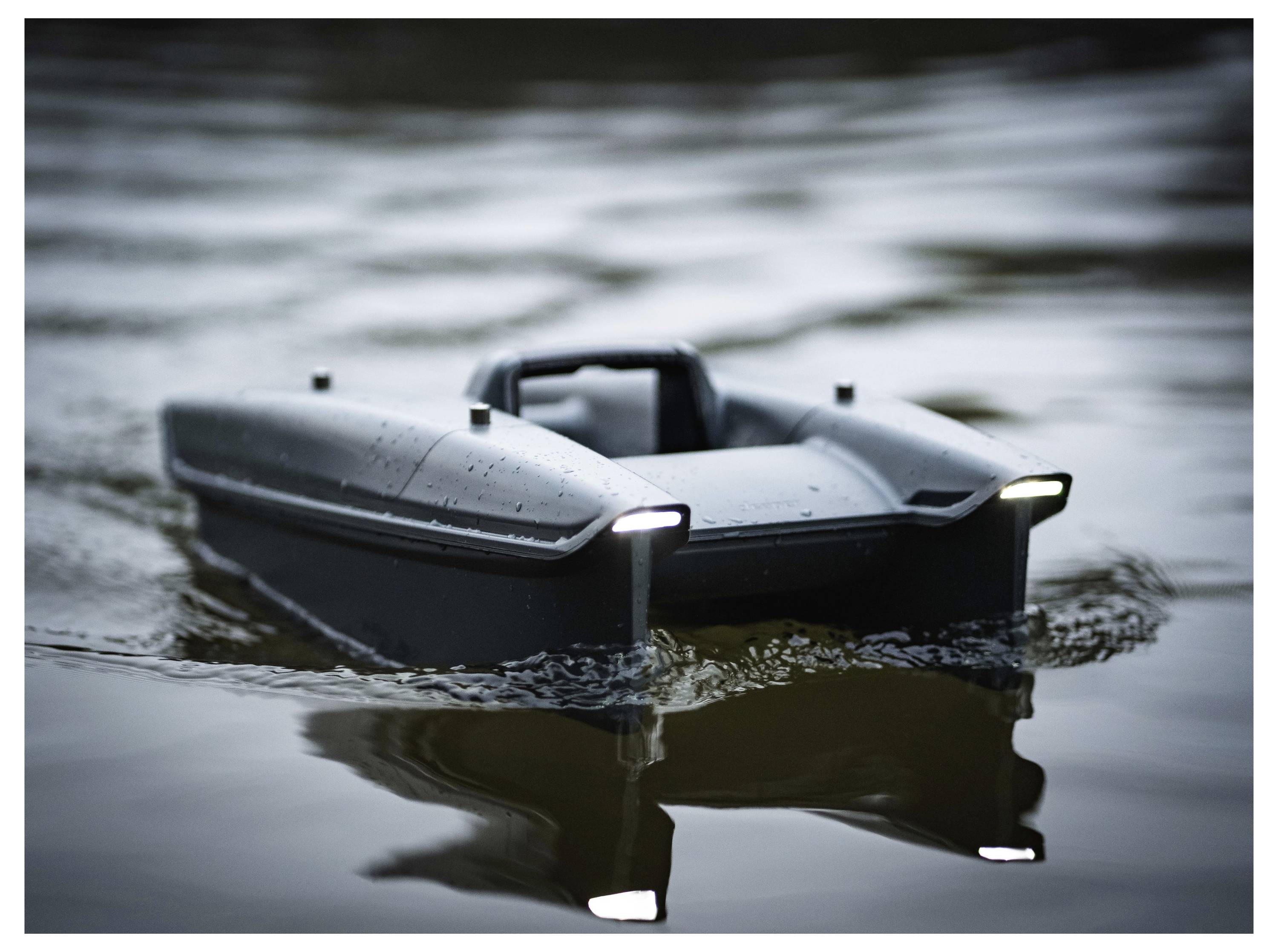 Un petit bateau autonome noir flotte sur une eau calme, avec des lumières allumées à l'avant et son reflet visible en dessous.