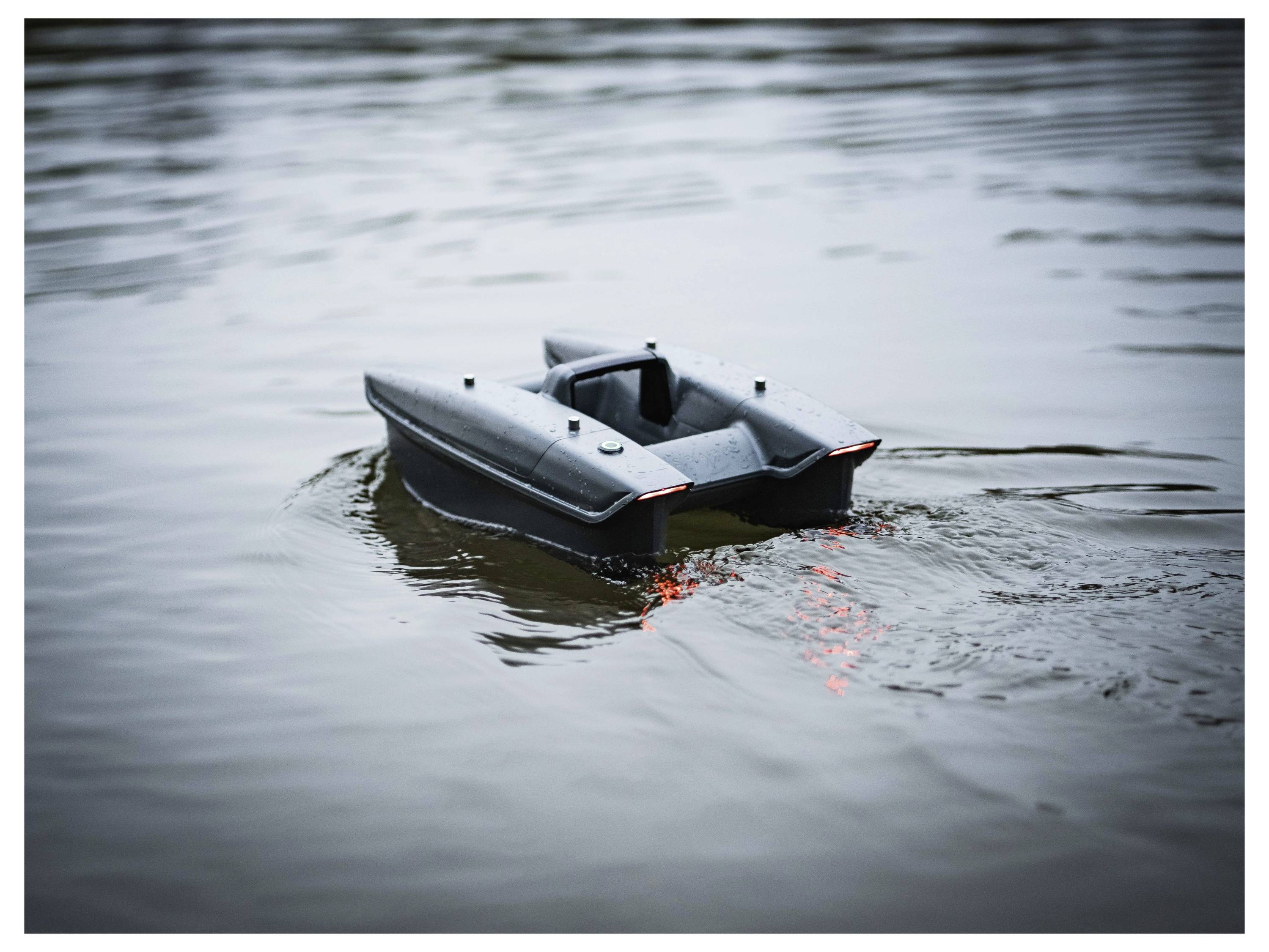 Un petit bateau radiocommandé noir flotte sur une eau calme, créant des ondulations et un léger sillage.