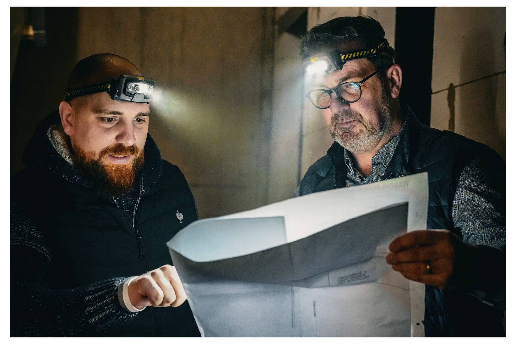 Deux hommes portant des lampes frontales étudient un grand plan dans une pièce faiblement éclairée, l'un pointant du doigt vers les plans.