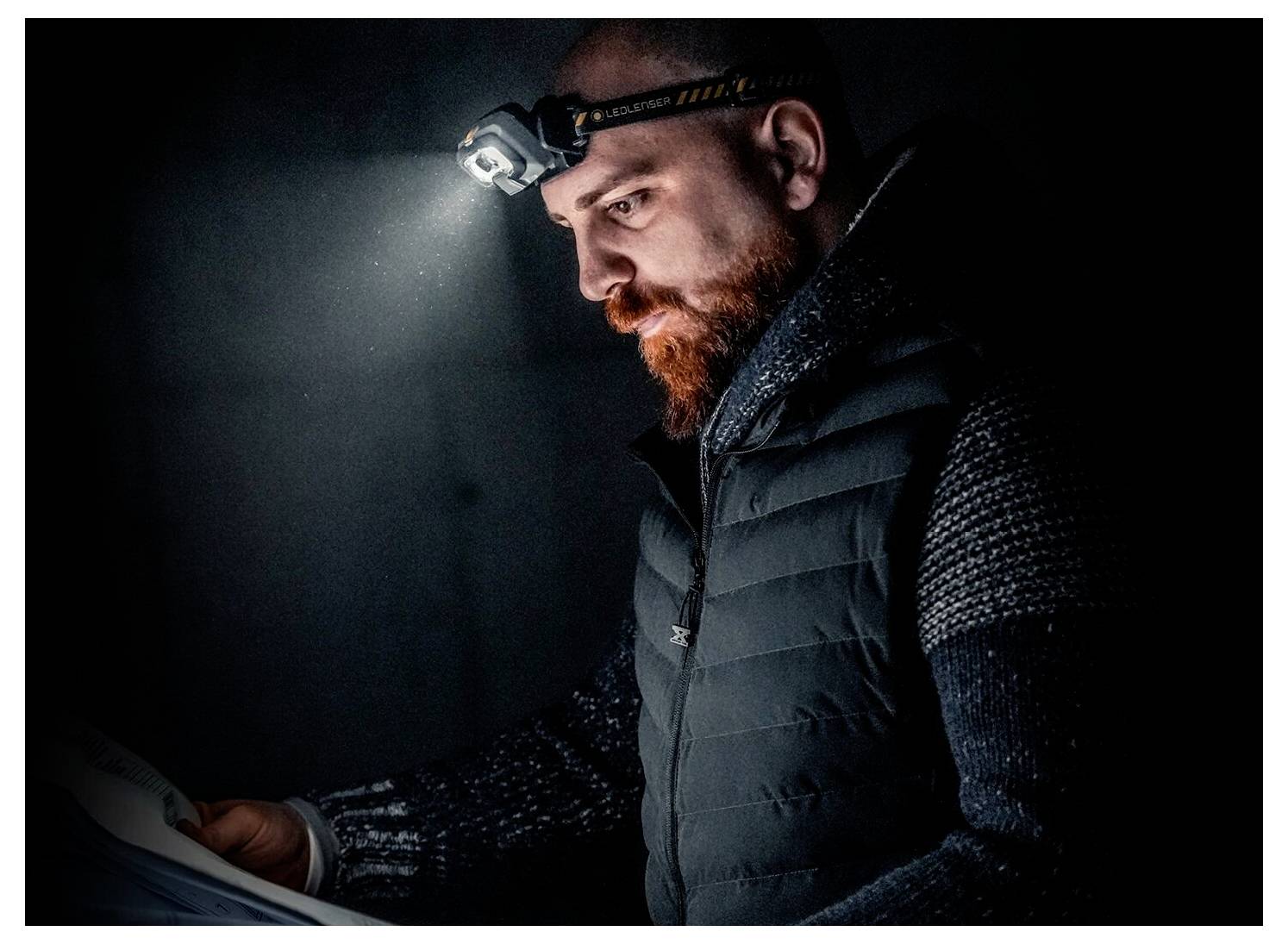 Homme barbu portant une lampe frontale et un gilet lit une carte dans un cadre extérieur sombre.