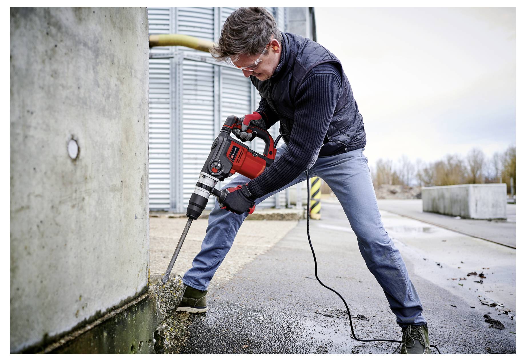 Un ouvrier utilise un marteau-piqueur pour briser du béton à côté d'un grand mur à l'extérieur sur un chantier de construction.