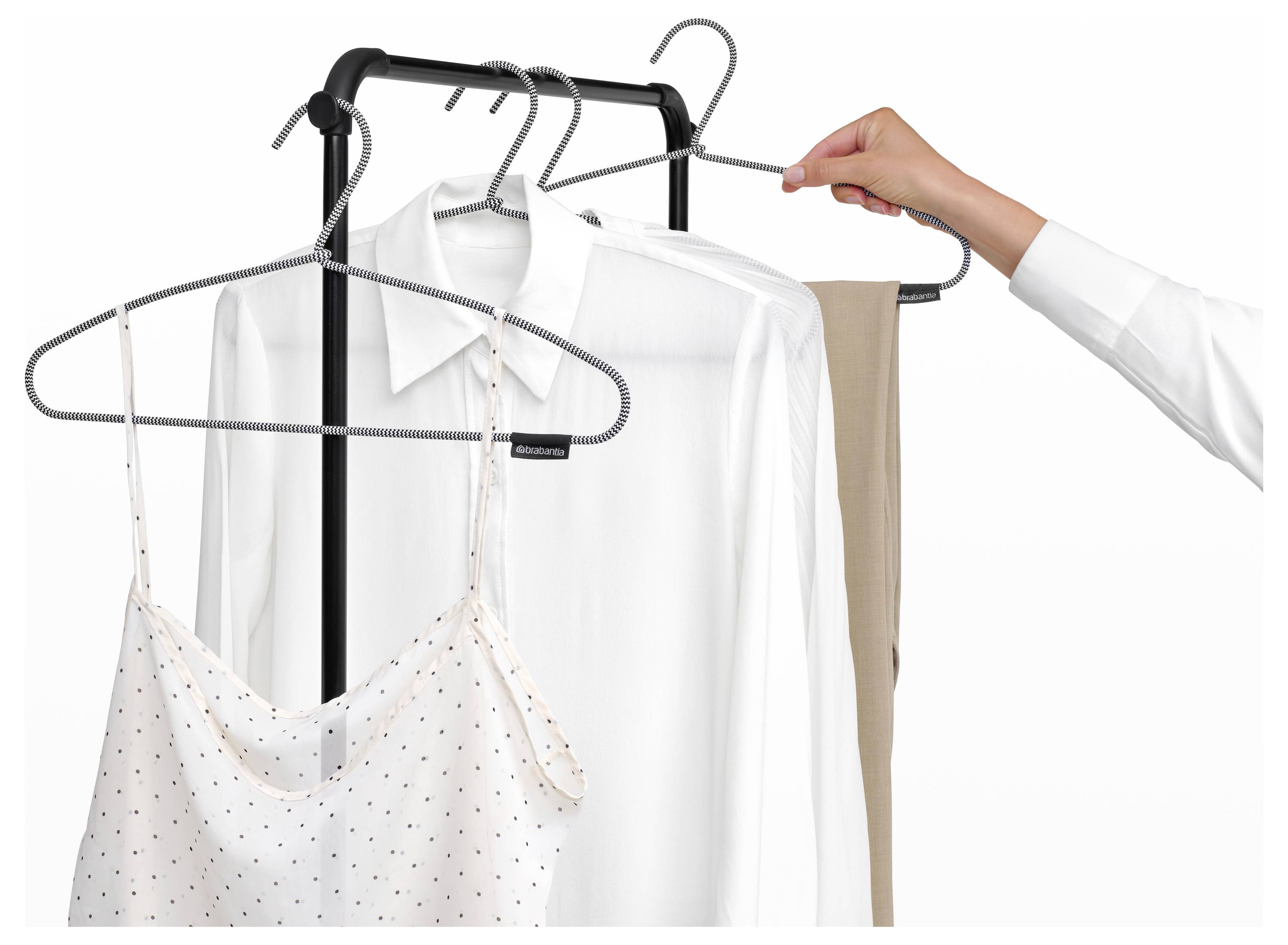 La main d'une personne suspend des chemises blanches, un vêtement beige et un chemisier à pois sur un portant à vêtements sur fond blanc.