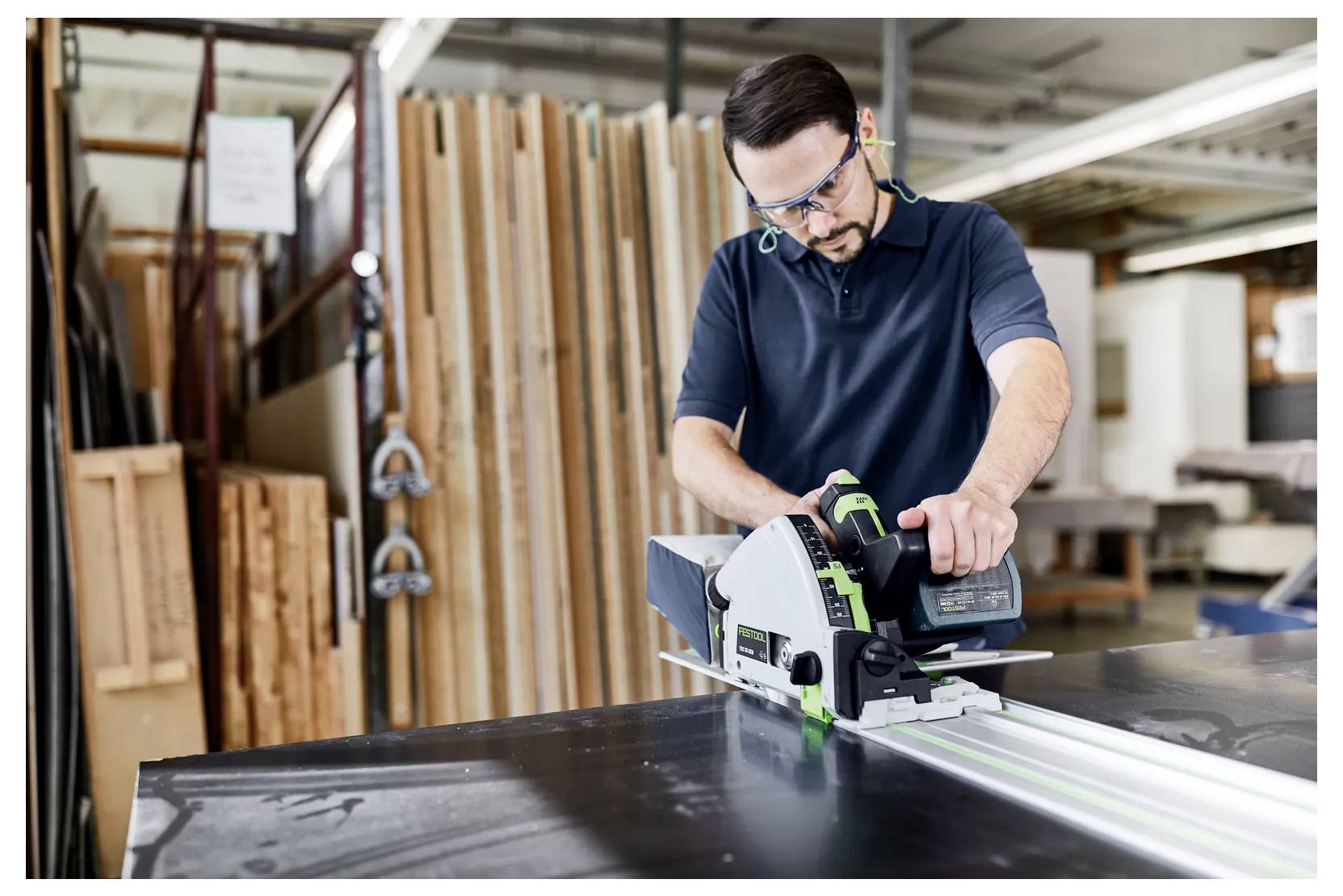 Menuisier utilisant une scie circulaire sur une grande planche dans un atelier, avec du bois empilé et des outils en arrière-plan.