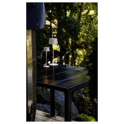 Deux petites lampes de table blanches sur une table noire d'extérieur sur un porche, avec des arbres ensoleillés et un jardin en arrière-plan.